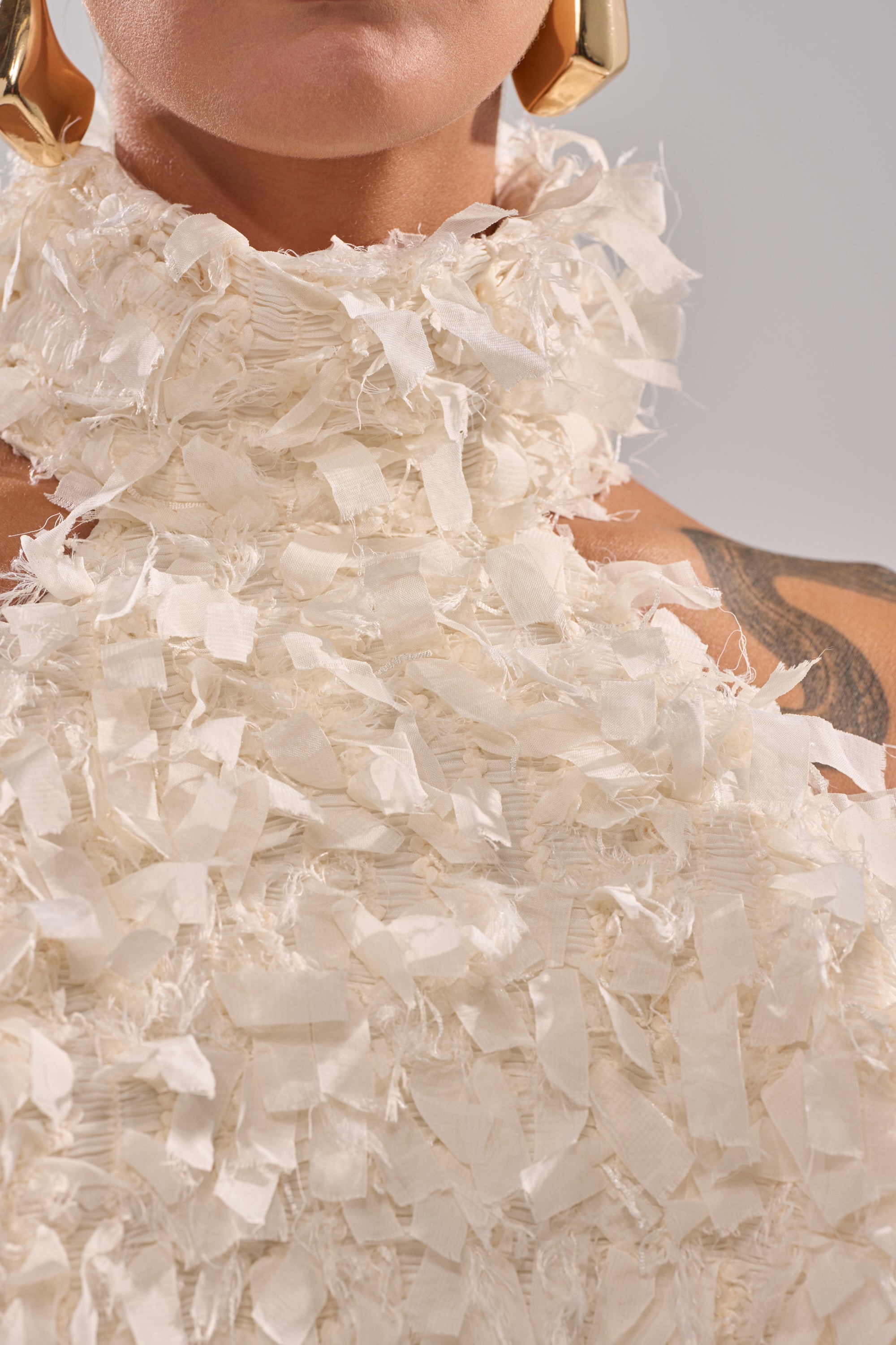 Close-up of a person wearing the MOXIE HALTER NECK BLOUSE, an off-white ruched mesh top with layered fringed fabric strips. Gold earrings and part of a tattoo are visible on the shoulder against a neutral background. 8
