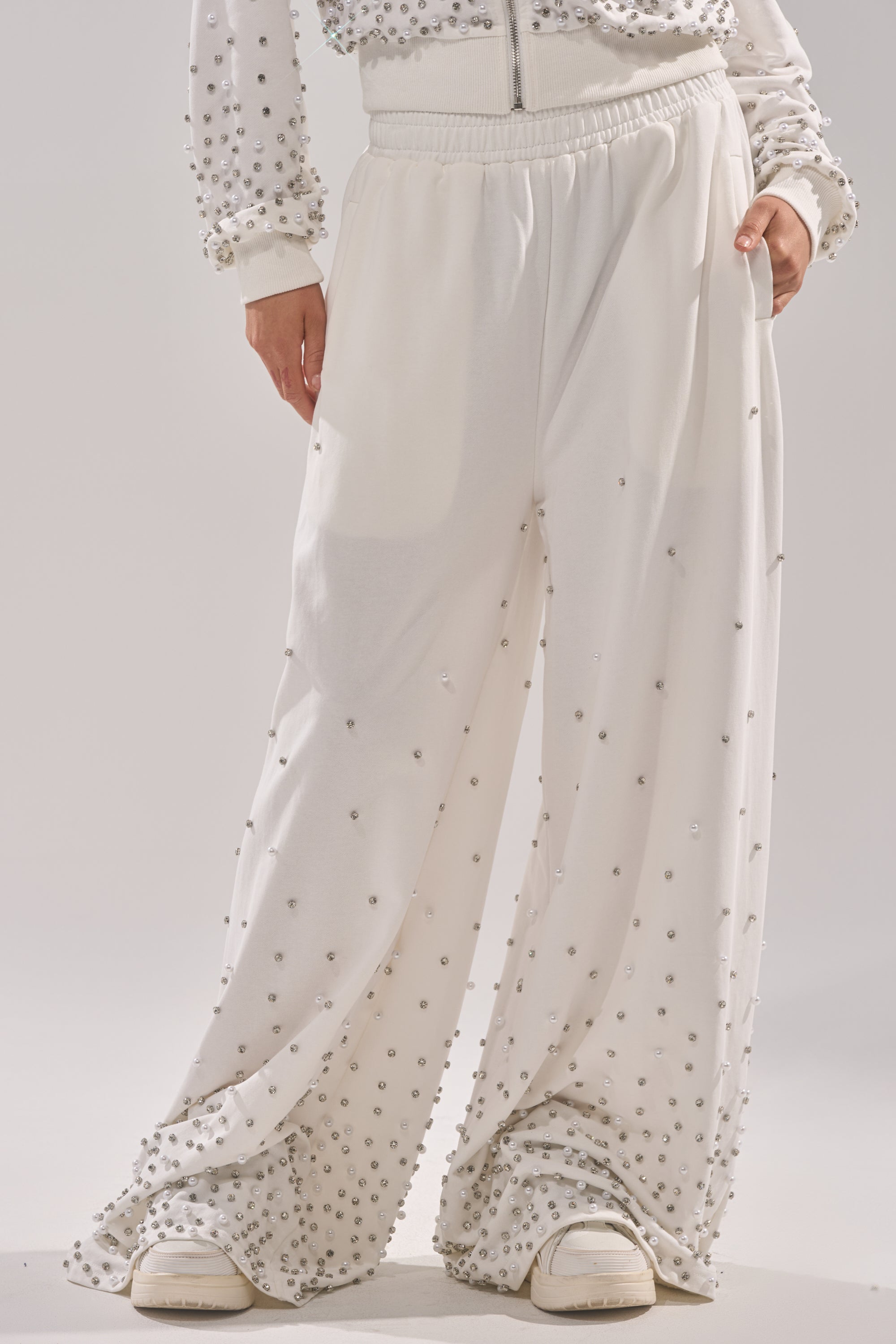 Someone stands against a plain background wearing the ALL IN WHITE RHINESTONE WIDE LEG JOGGER with white sneakers and a matching rhinestone-studded jacket. 1