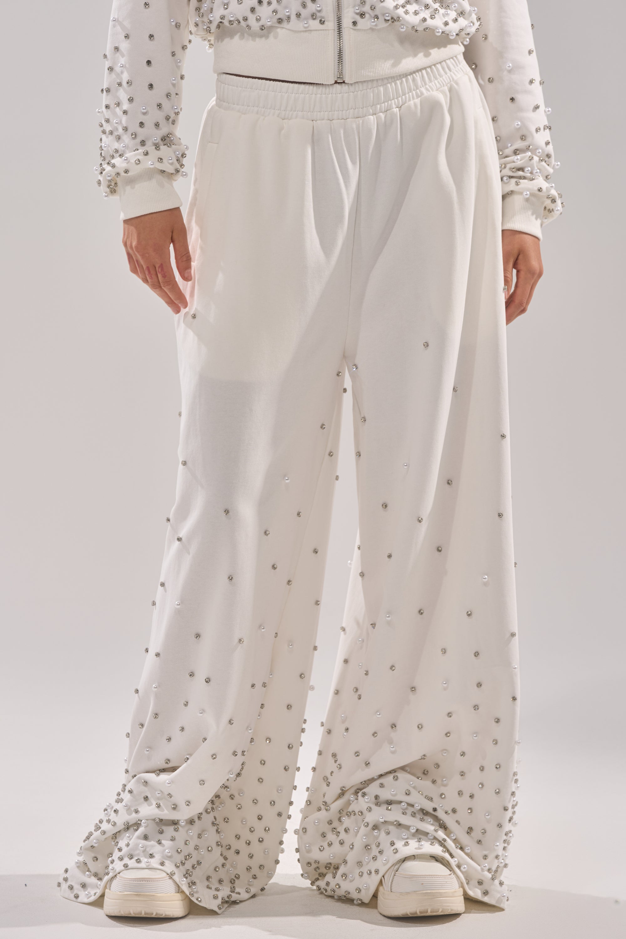 A person wearing the ALL IN WHITE RHINESTONE WIDE LEG JOGGER, featuring loose-fitting white joggers adorned with silver studs, paired with white sneakers and a matching studded white jacket. 2