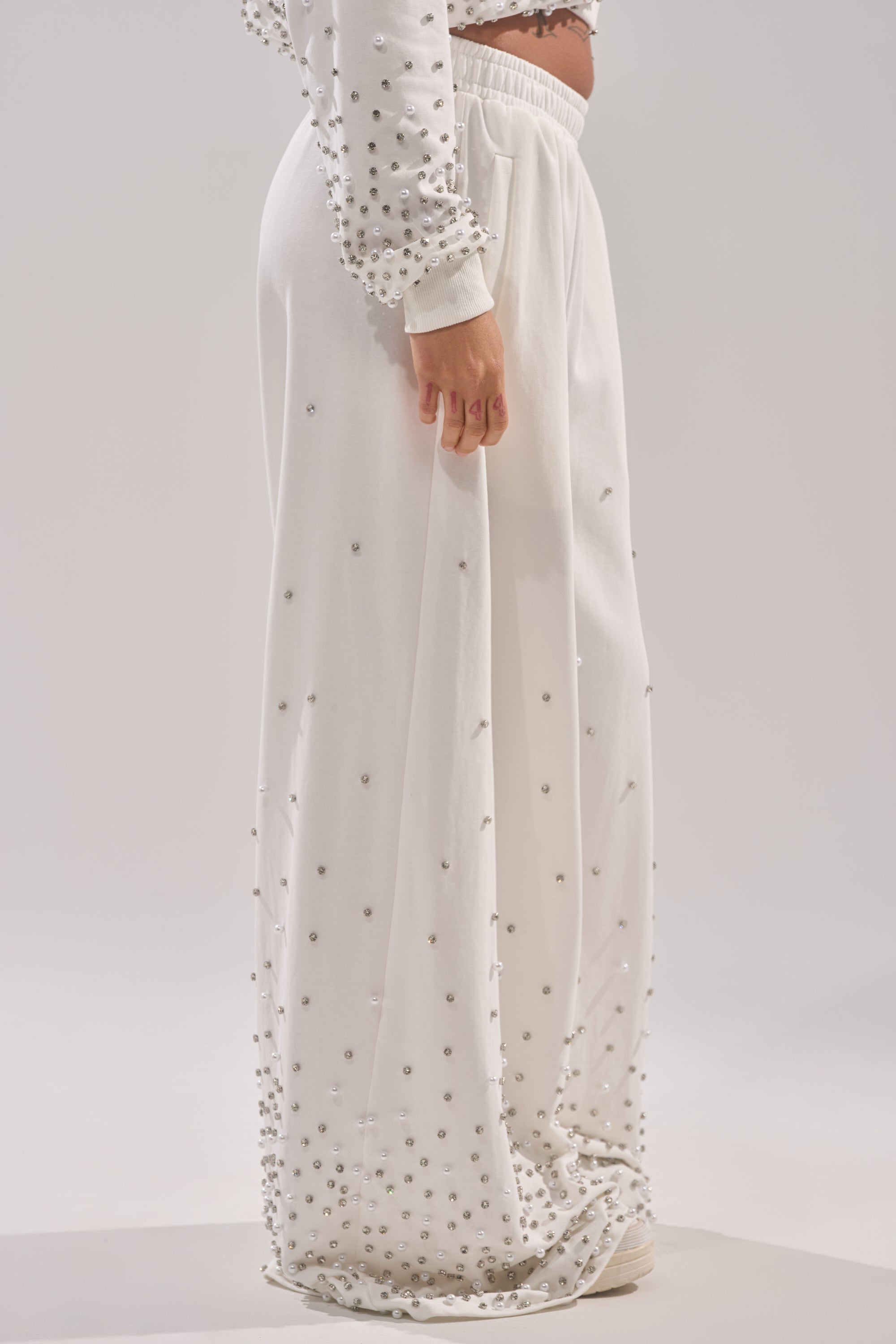 A person wearing the ALL IN WHITE RHINESTONE WIDE LEG JOGGER and a matching top, both decorated with small silver studs. The photo, taken from the side, highlights the lower body against a neutral background. 3