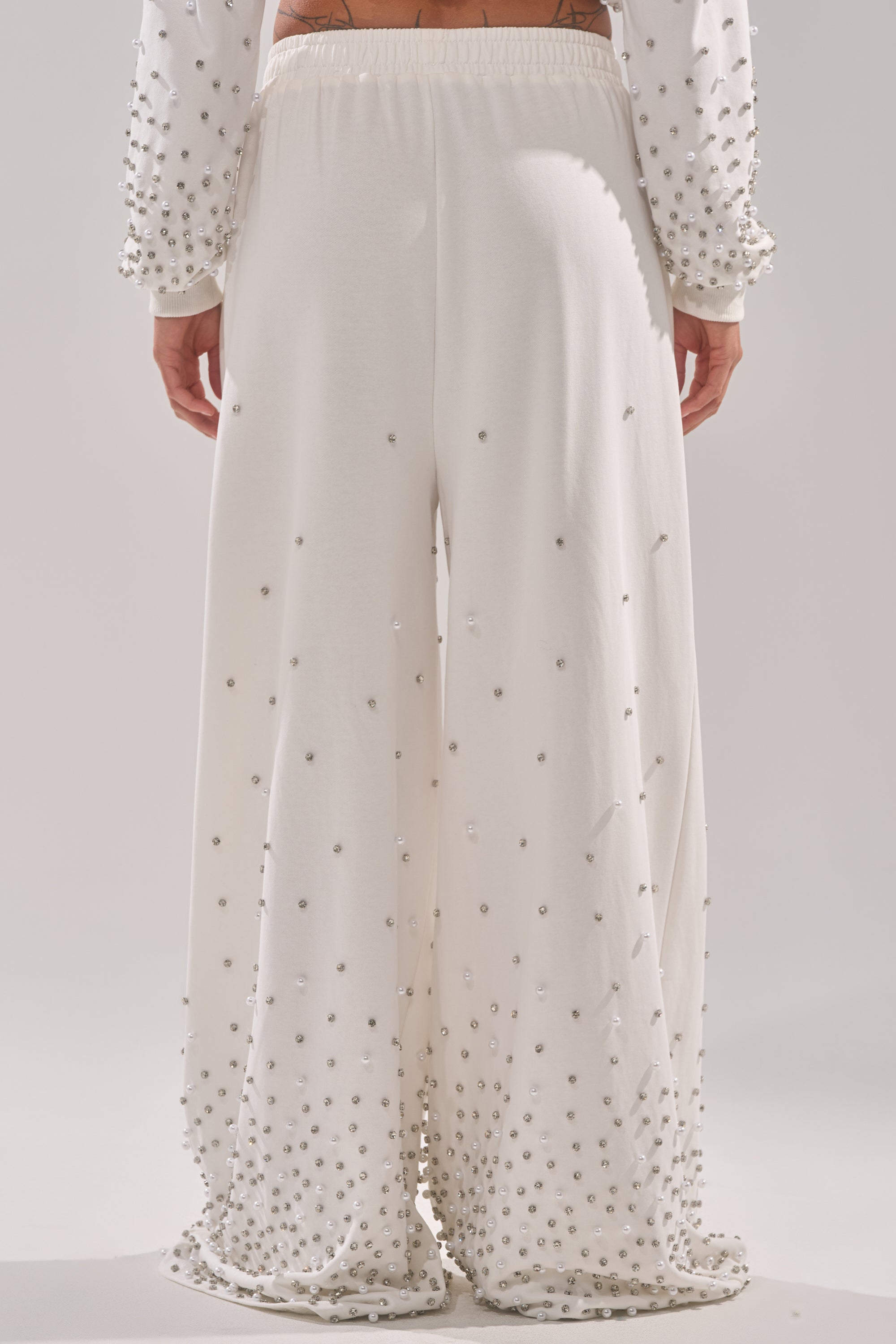 A person shown from the back wears the ALL IN WHITE RHINESTONE WIDE LEG JOGGER—loose-fitting white pants with small round silver embellishments concentrated at the bottom and spreading out toward the top. 4