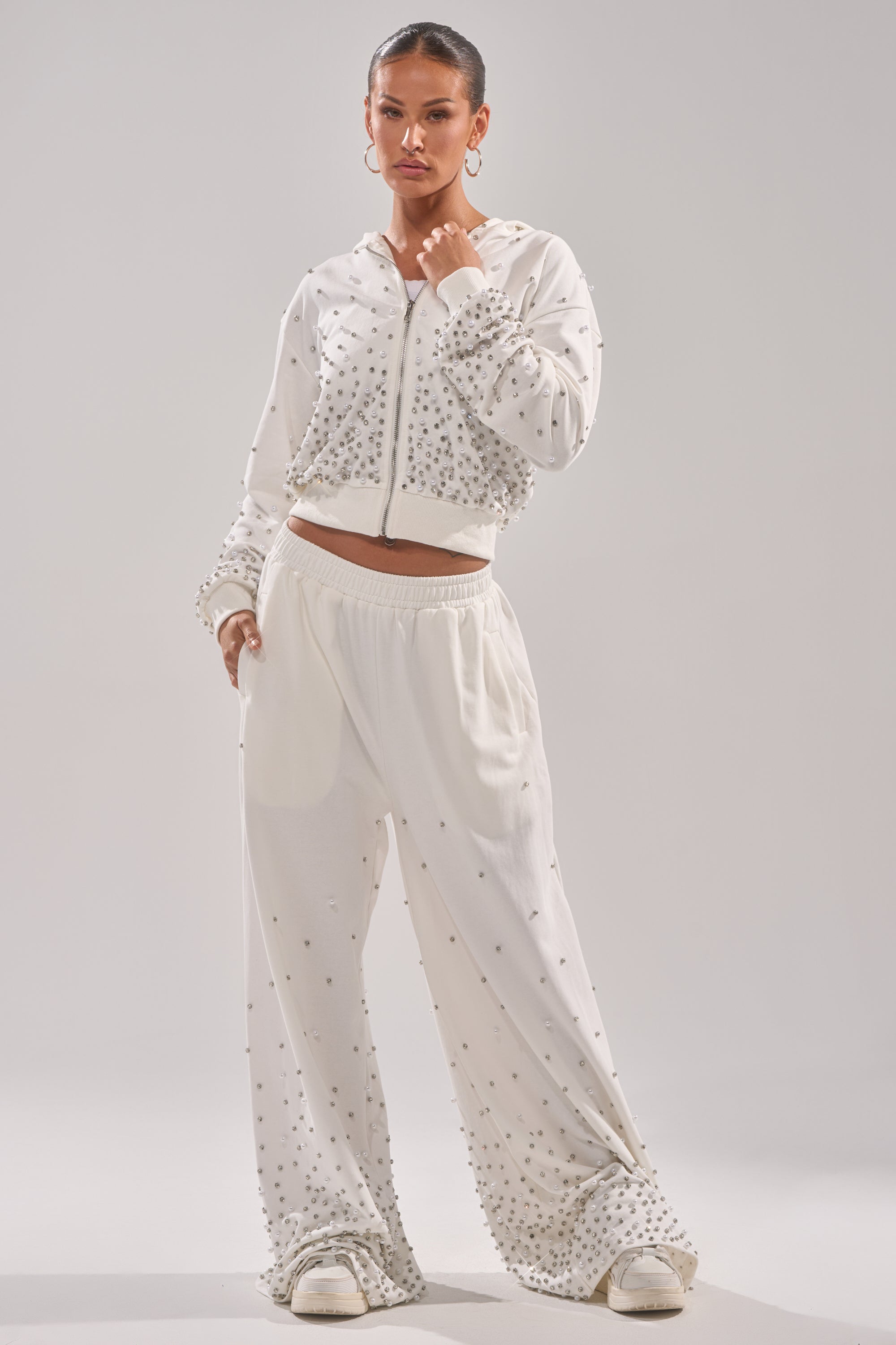 A woman poses in ALL IN WHITE RHINESTONE WIDE LEG JOGGERS and a white hoodie, styled with hoop earrings and white sneakers against a plain light background. 5
