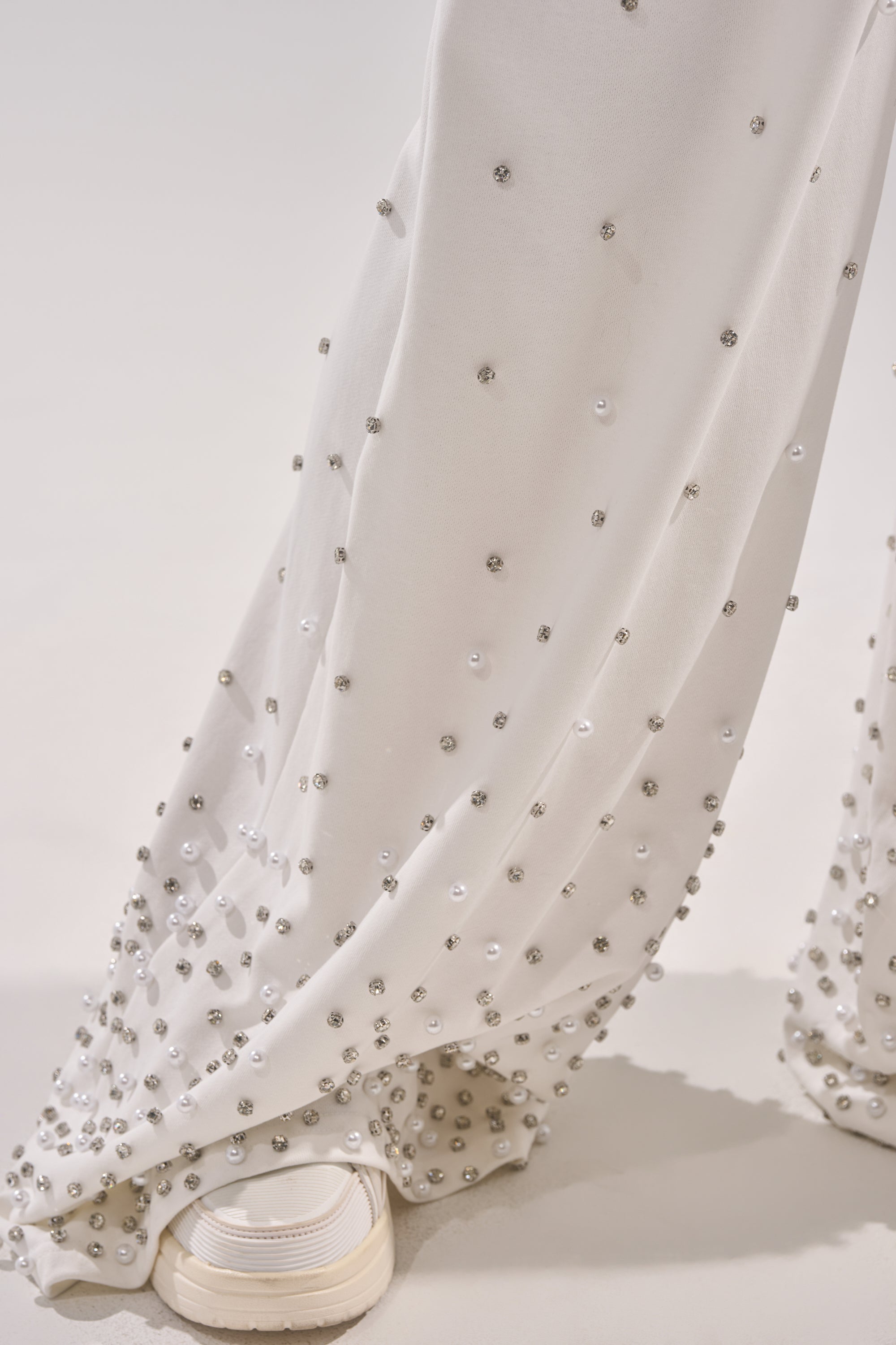 Close-up of the ALL IN WHITE RHINESTONE WIDE LEG JOGGER featuring small, shiny beads and pearls, draped over white sneakers against a plain light background. 6