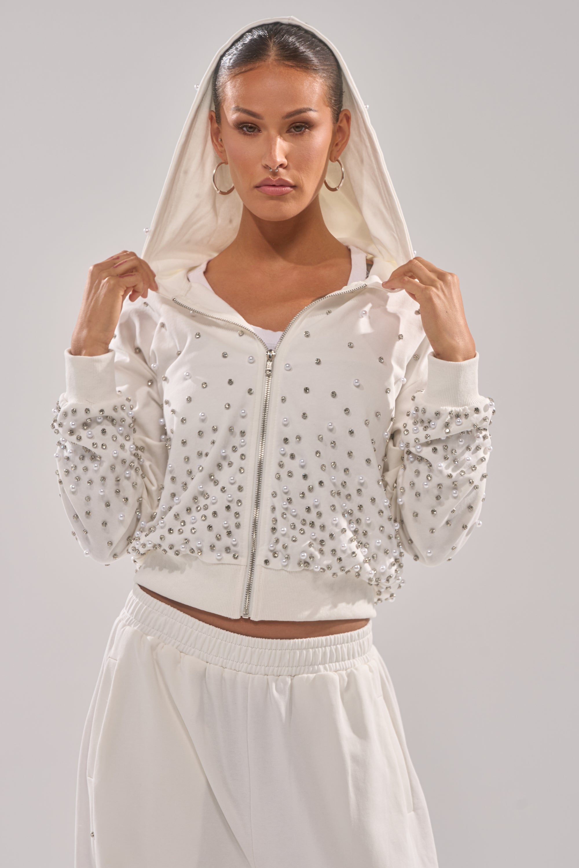 A woman models the ALL IN WHITE RHINESTONE ZIP UP SWEATSHIRT, featuring pearl embellishments and a hood, paired with white pants against a plain light background. She holds the hoodie edges near her face, showcasing confident loungewear style. 1