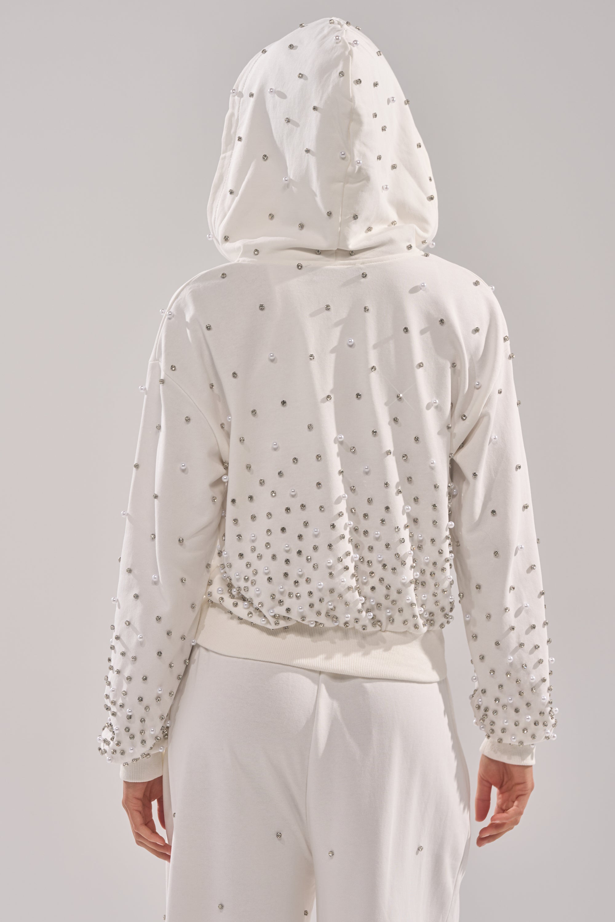 Someone wears the ALL IN WHITE RHINESTONE ZIP UP SWEATSHIRT, featuring clusters of silver studs on the back and sleeves, paired with white pants. The stylish look is photographed from behind against a light background. 3