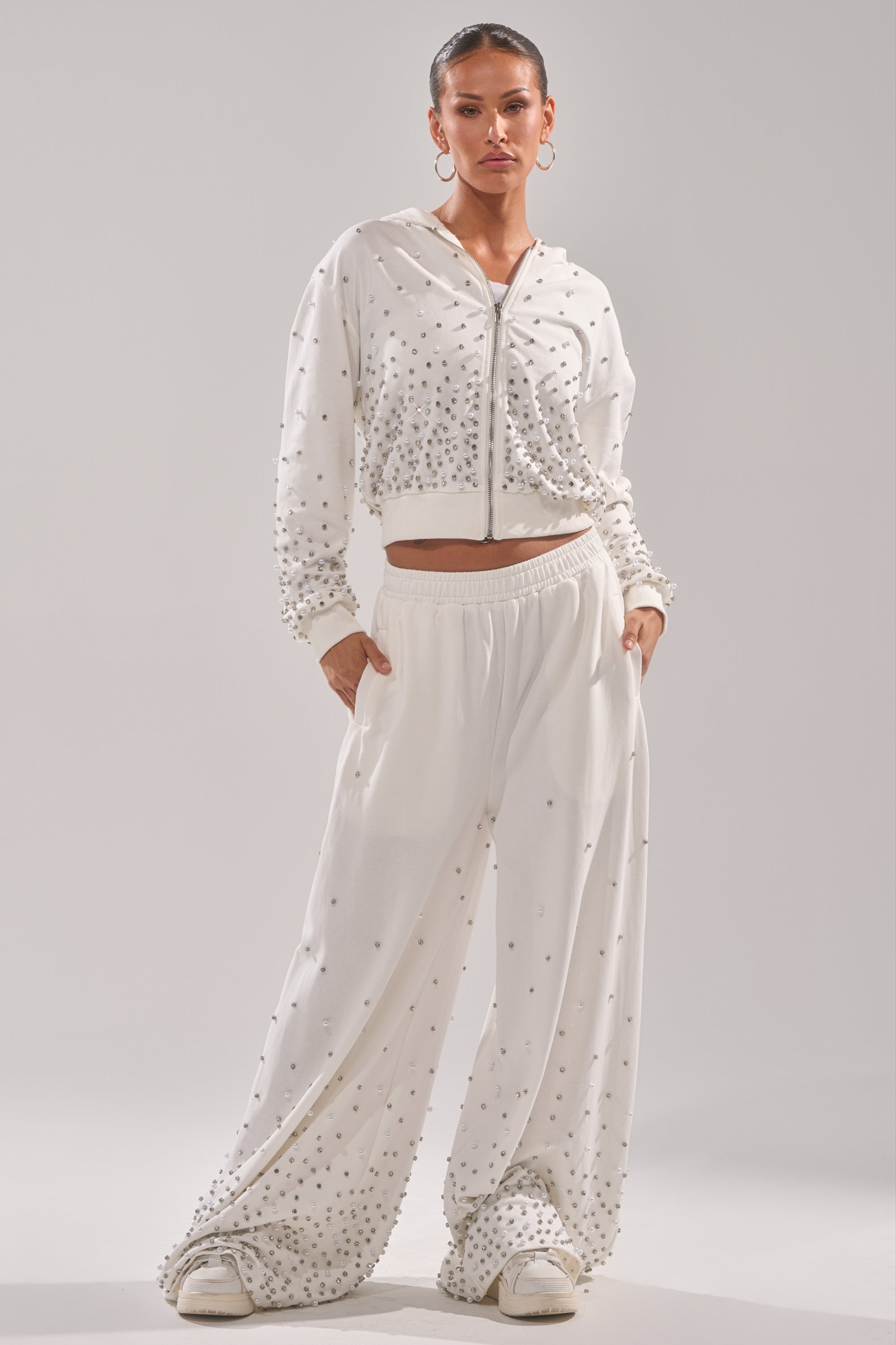 A person stands confidently in the ALL IN WHITE RHINESTONE ZIP UP SWEATSHIRT and matching wide-leg pants, both with pearl embellishments. Hoop earrings and white shoes complete the chic look, hands casually in pockets. 4