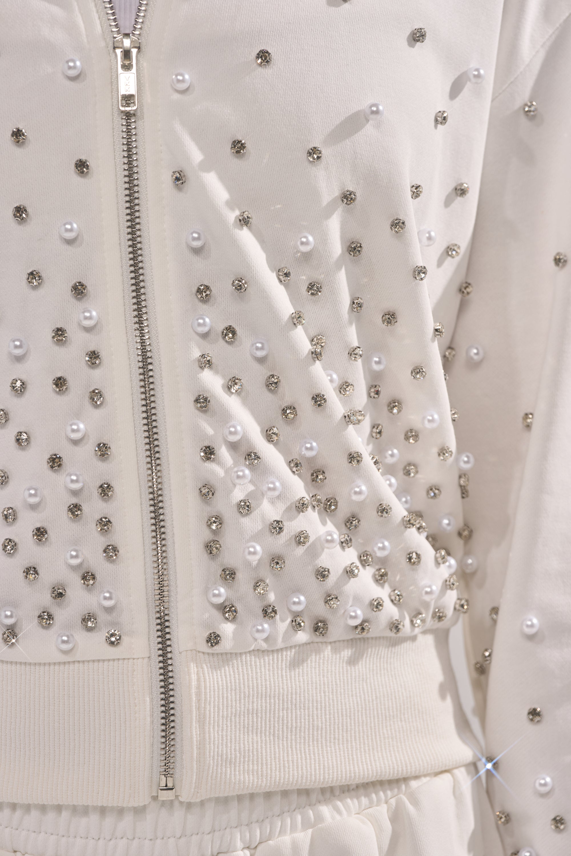 The ALL IN WHITE RHINESTONE ZIP UP SWEATSHIRT features clusters of silver studs and pearl embellishments on white fabric, creating a textured, sparkly look that's perfect for stylish loungewear. 6