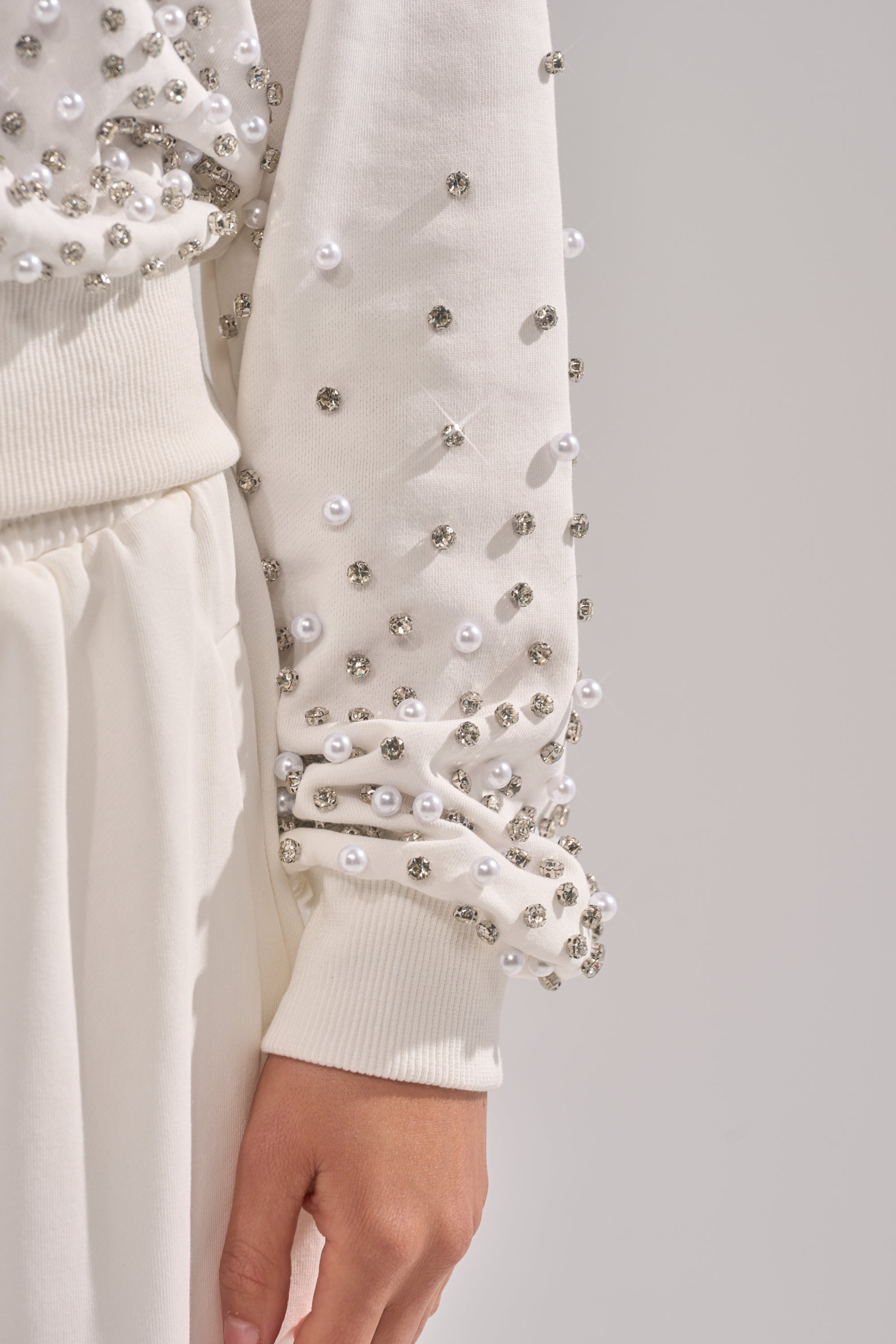 A close-up of a person wearing the ALL IN WHITE RHINESTONE ZIP UP SWEATSHIRT with pearl details, paired with matching white loungewear pants. Their arm rests by their side against a neutral background. 7