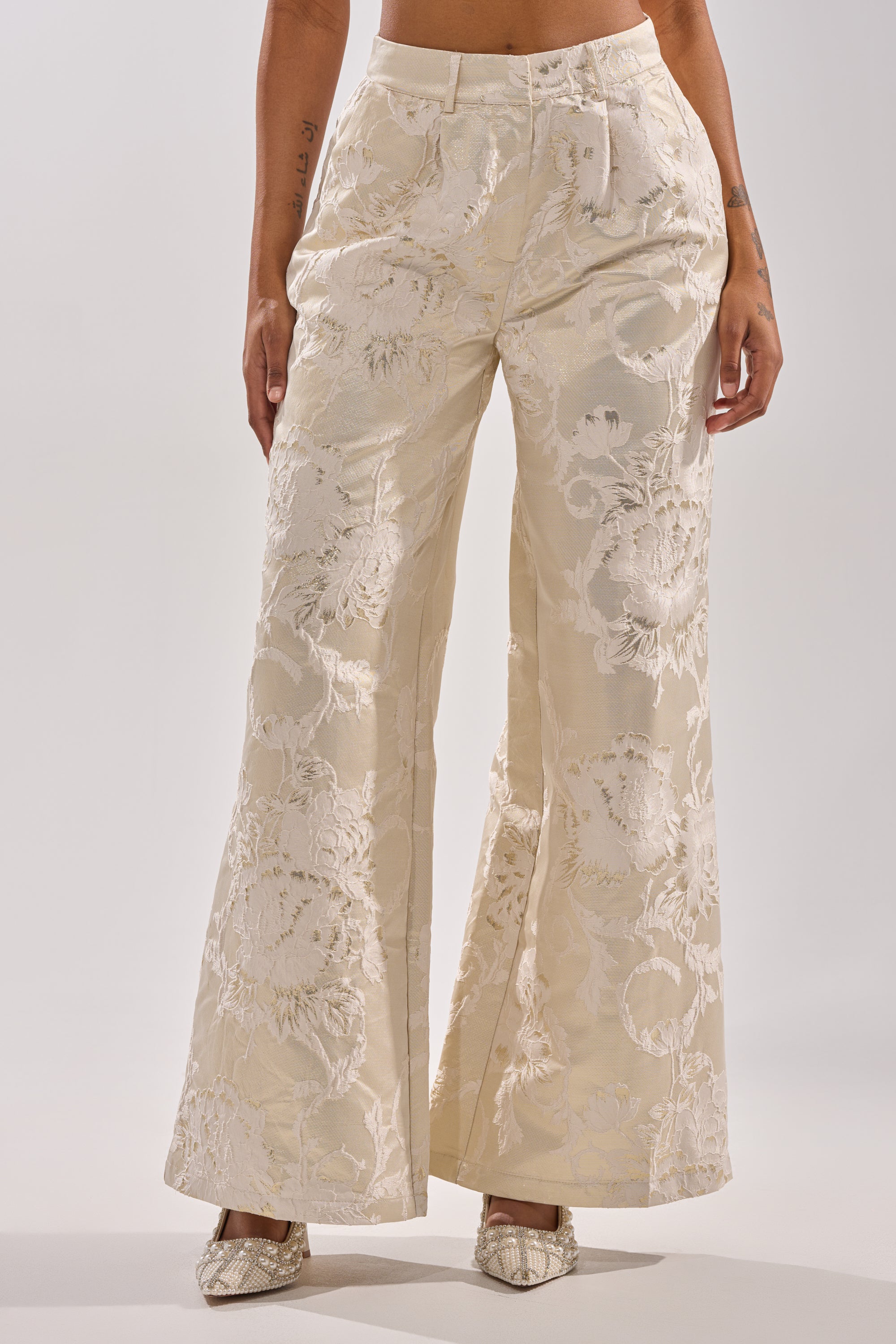 A person wears the DRESSED TO IMPRESS TROUSER, featuring cream-colored jacquard fabric with a floral pattern and wide legs, styled with pointed-toe shoes against a plain white background. 0