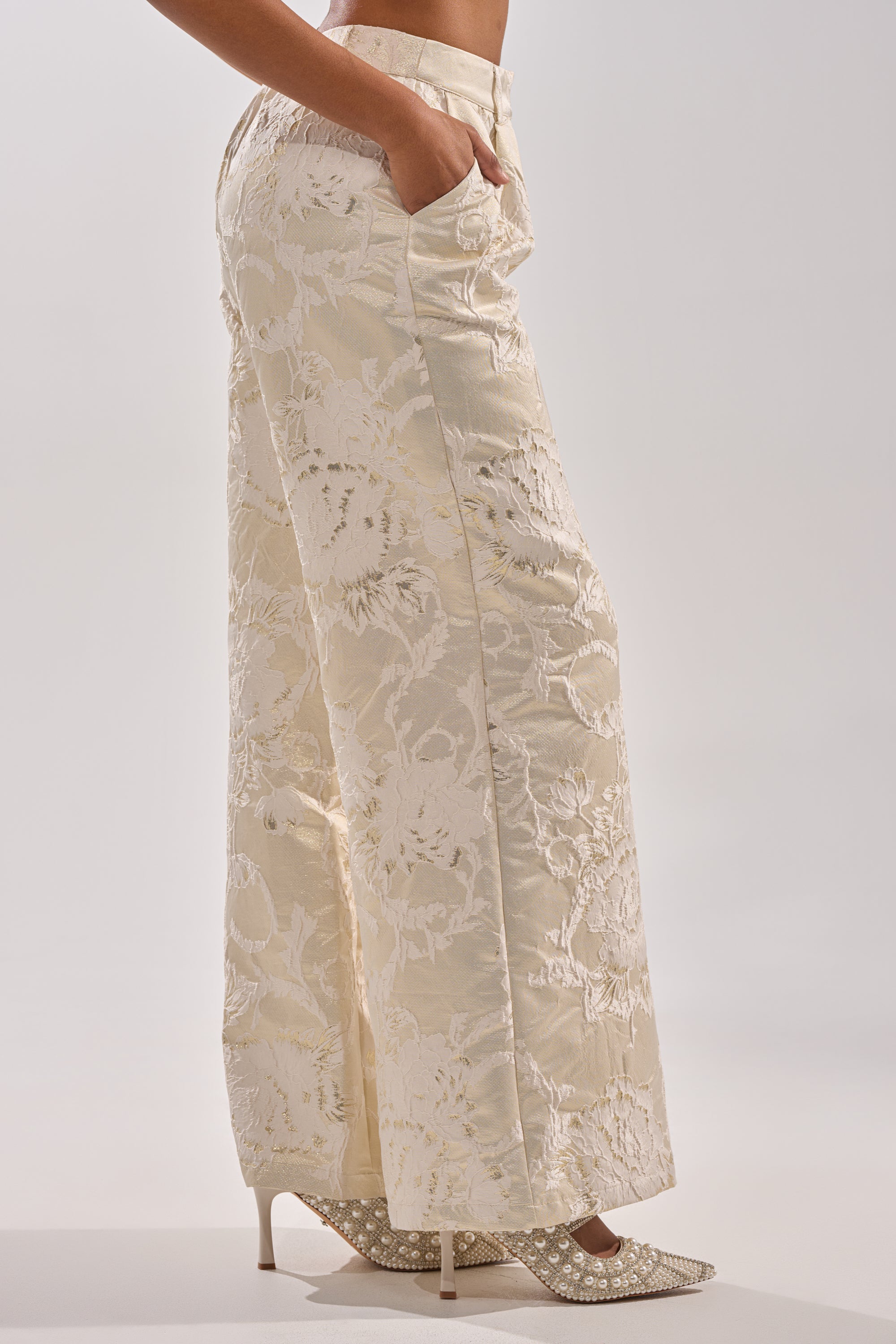 Wearing the DRESSED TO IMPRESS TROUSER—high-rise, wide-leg cream brocade pants with a floral pattern—paired with embellished cream heels, hands in pockets, stands against a plain light background. 1