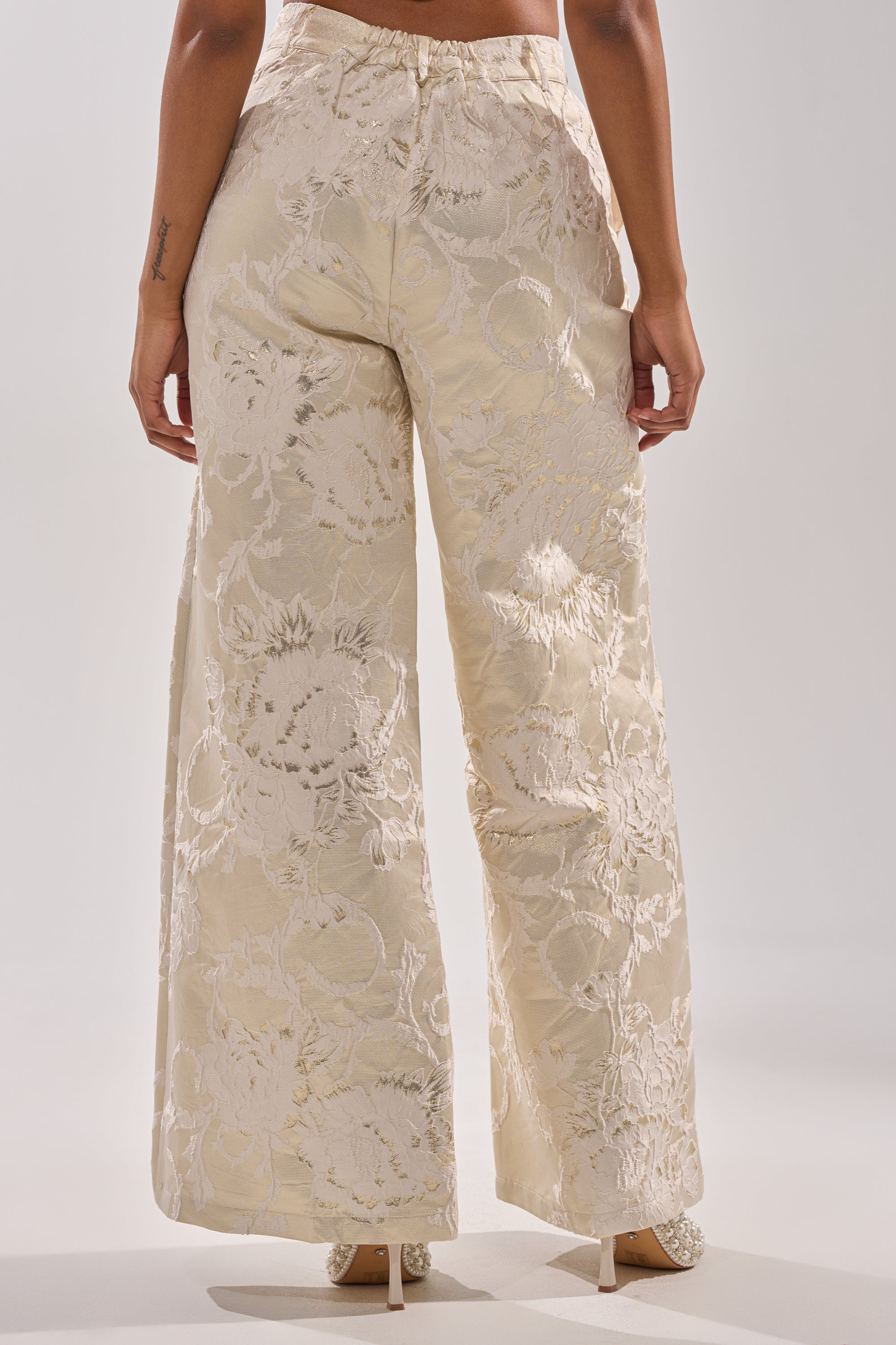 A person faces away, wearing the DRESSED TO IMPRESS TROUSER by AKIRA Label, featuring a high-rise wide-leg fit with a pearlescent jacquard pattern and paired with white heels against a plain light backdrop. 2