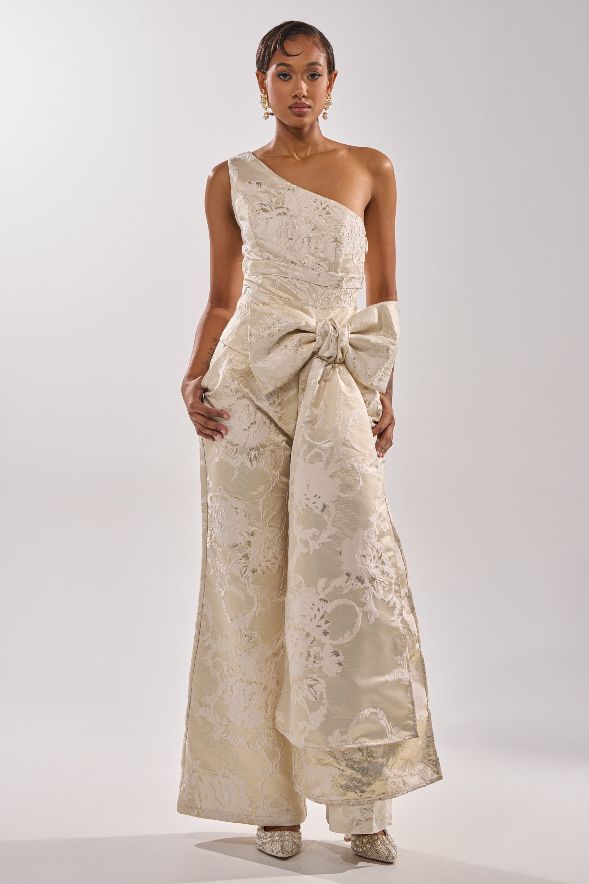 A woman wears the DRESSED TO IMPRESS TROUSER—a cream, one-shoulder jumpsuit with a waist bow and wide-leg floral silhouette—paired with pointed shoes, set against a plain light background. 3