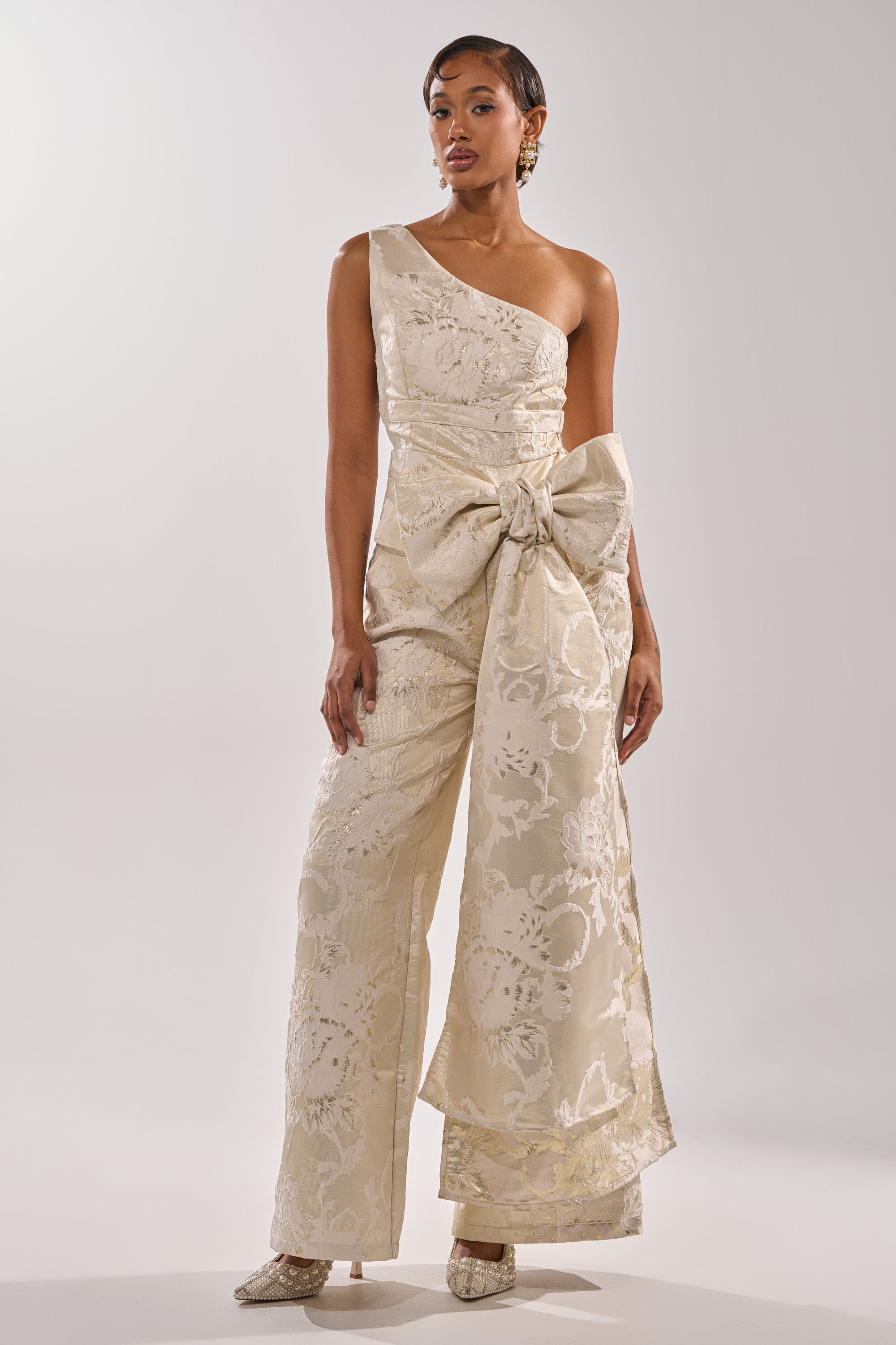 A woman stands against a plain background wearing the DRESSED TO IMPRESS TROUSER, a one-shoulder, cream-colored brocade jumpsuit with wide-leg silhouette and a dramatic waist bow, paired with matching heeled shoes. 4