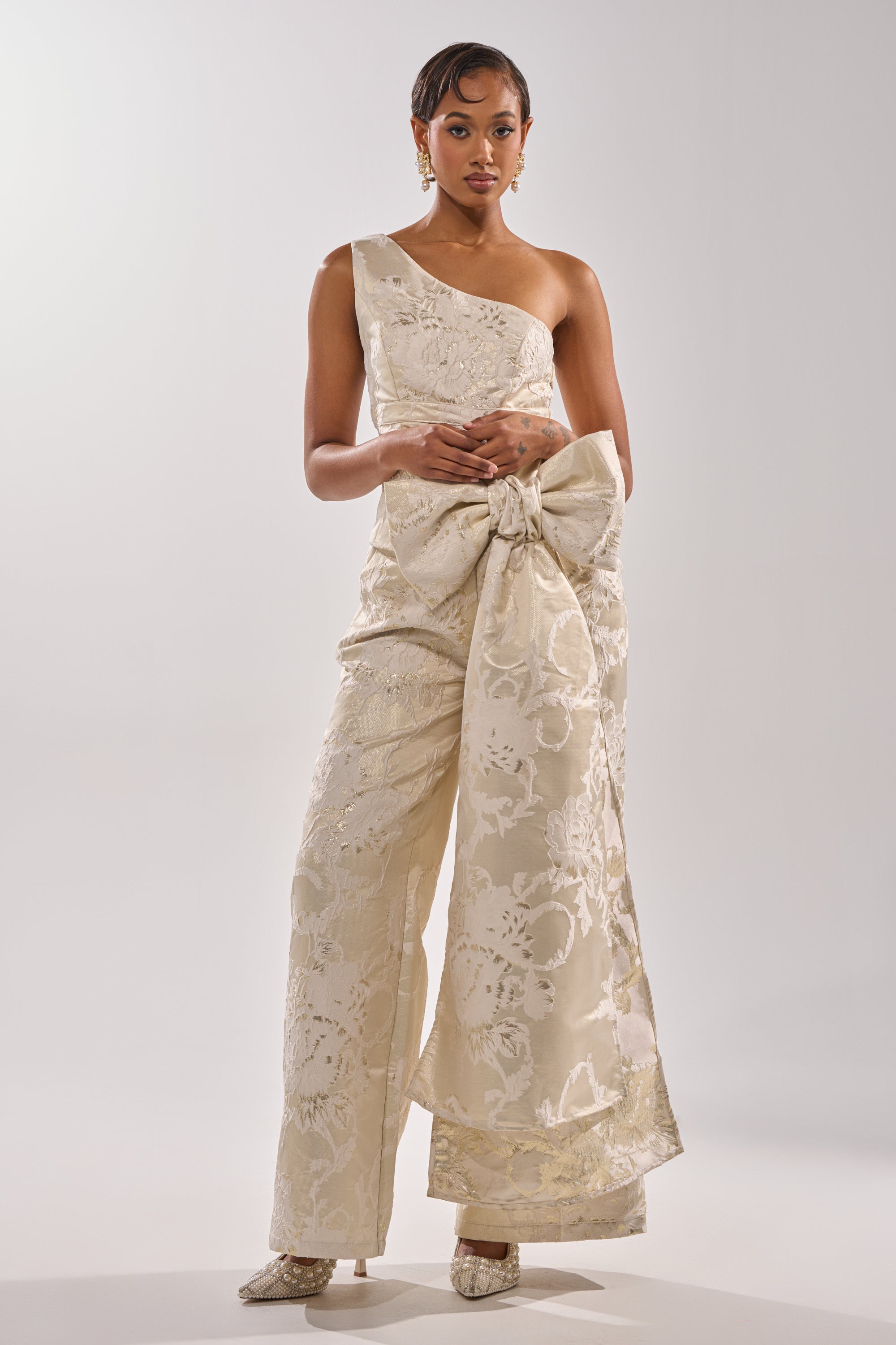 A woman wears the DRESSED TO IMPRESS TROUSER, an elegant cream one-shoulder jumpsuit featuring a wide-leg fit, a statement waist bow, and intricate floral patterns, styled with sleek hair, earrings, and pointed shoes. 5