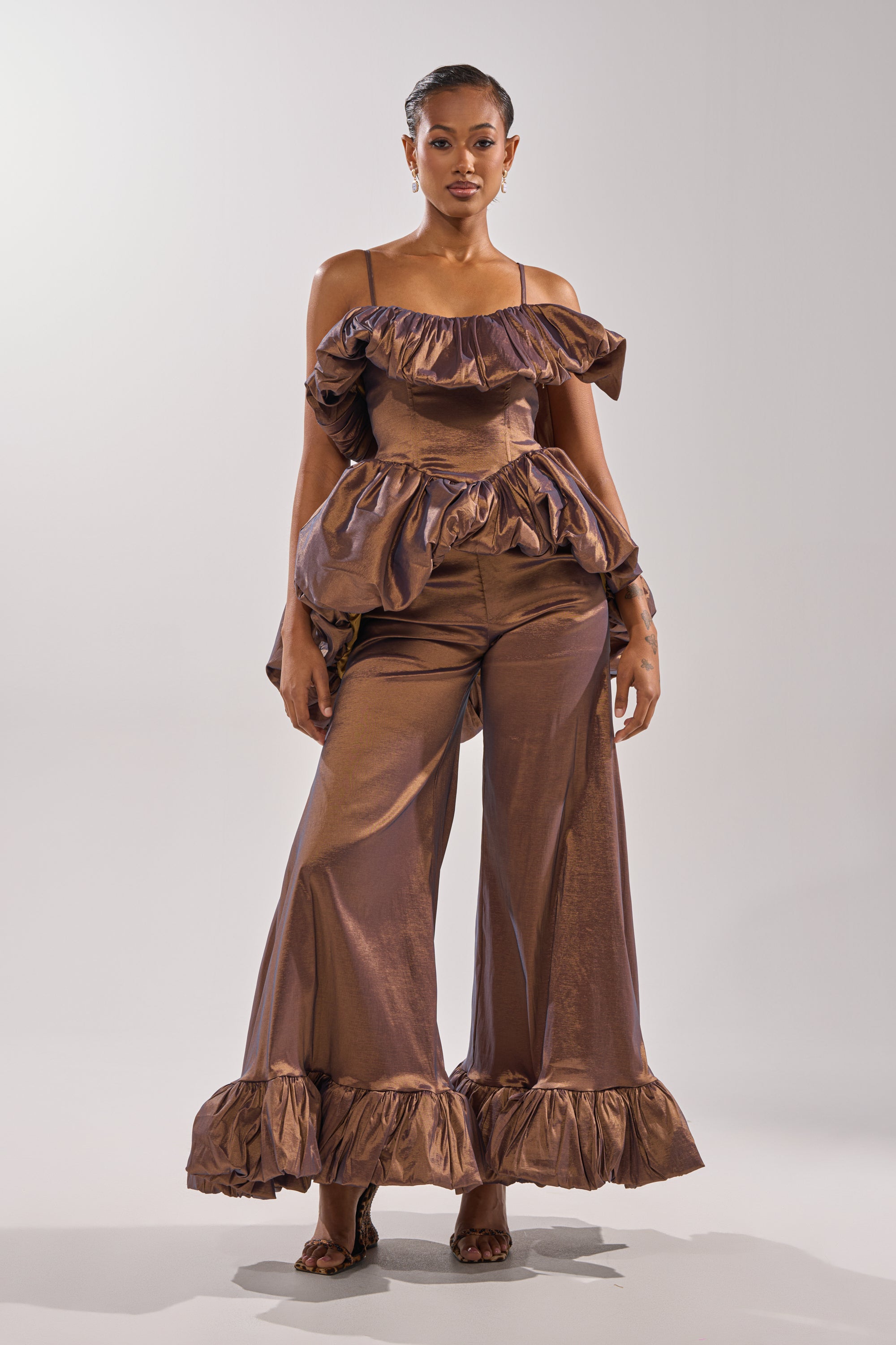 A woman stands confidently in the NOT TOO SIMPLE TROUSER, a shiny bronze high-rise metallic taffeta wide-leg pant with ruffled hems, paired with a matching ruffled off-shoulder top against a plain light background.