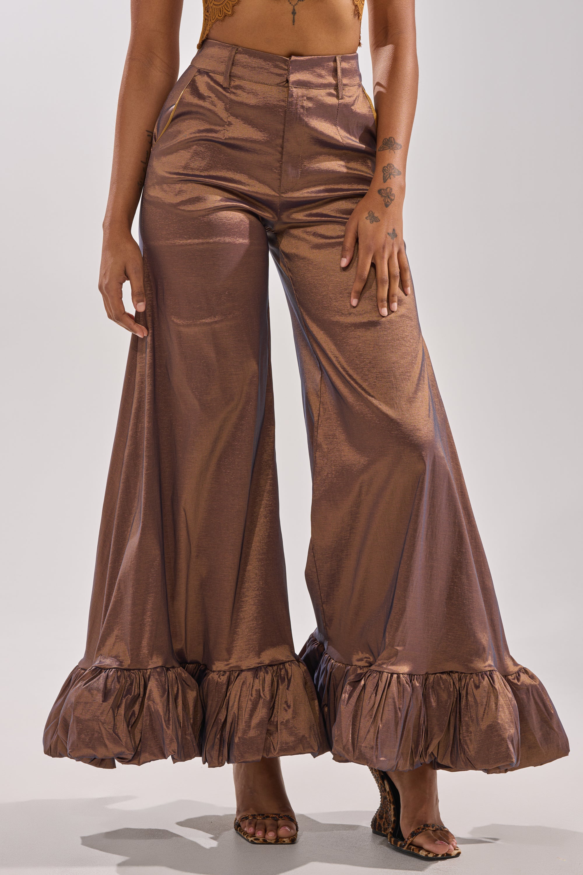 A person stands barefoot on a light background wearing the NOT TOO SIMPLE TROUSER, shiny bronze metallic taffeta wide-leg pants with ruffled hems. Their midriff is visible and their hands rest at their sides.