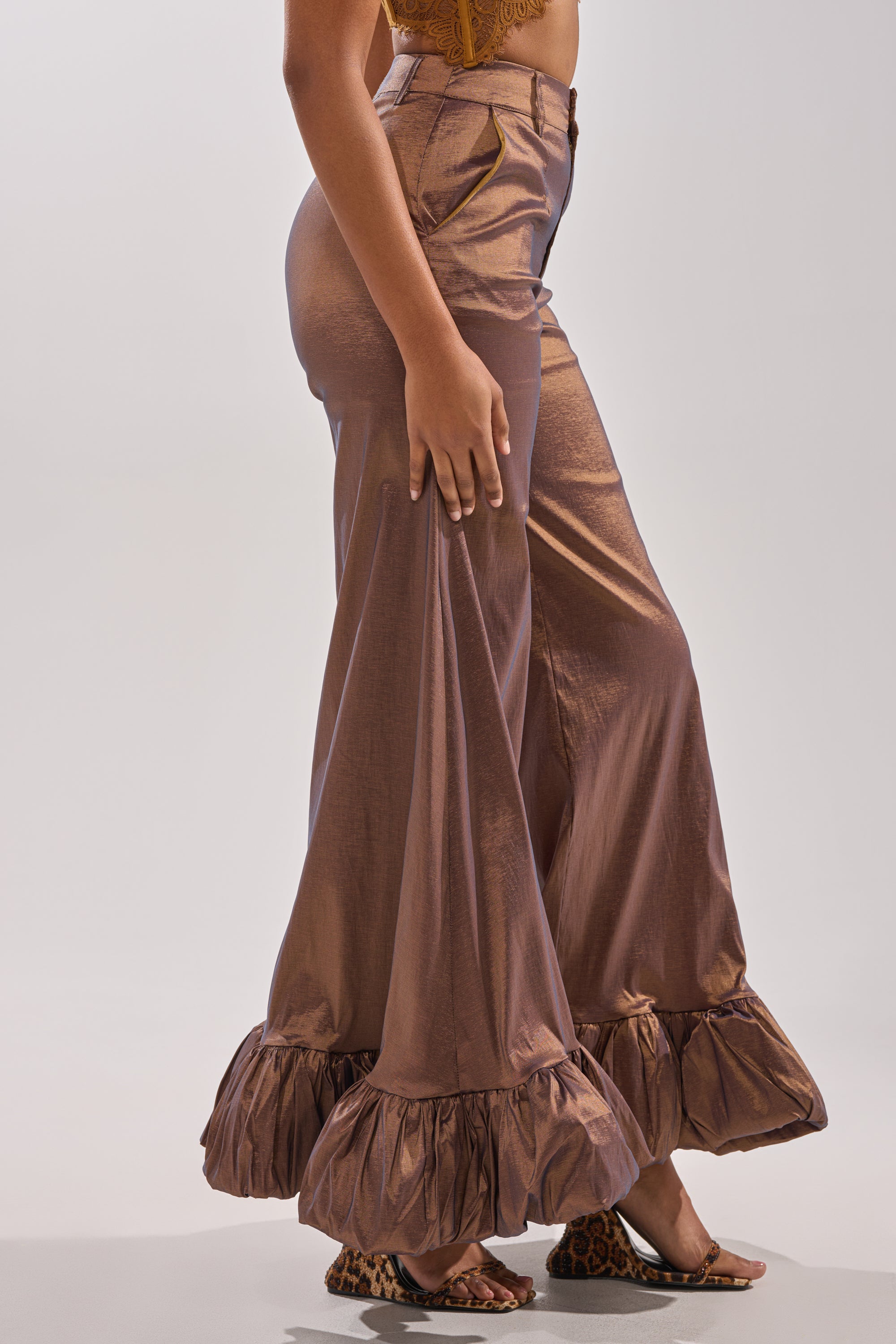 A person wears the NOT TOO SIMPLE TROUSER—shiny bronze metallic taffeta with a high-rise, ruffled bell-bottoms, and wide-leg silhouette—paired with leopard-print heeled sandals against a plain light background.