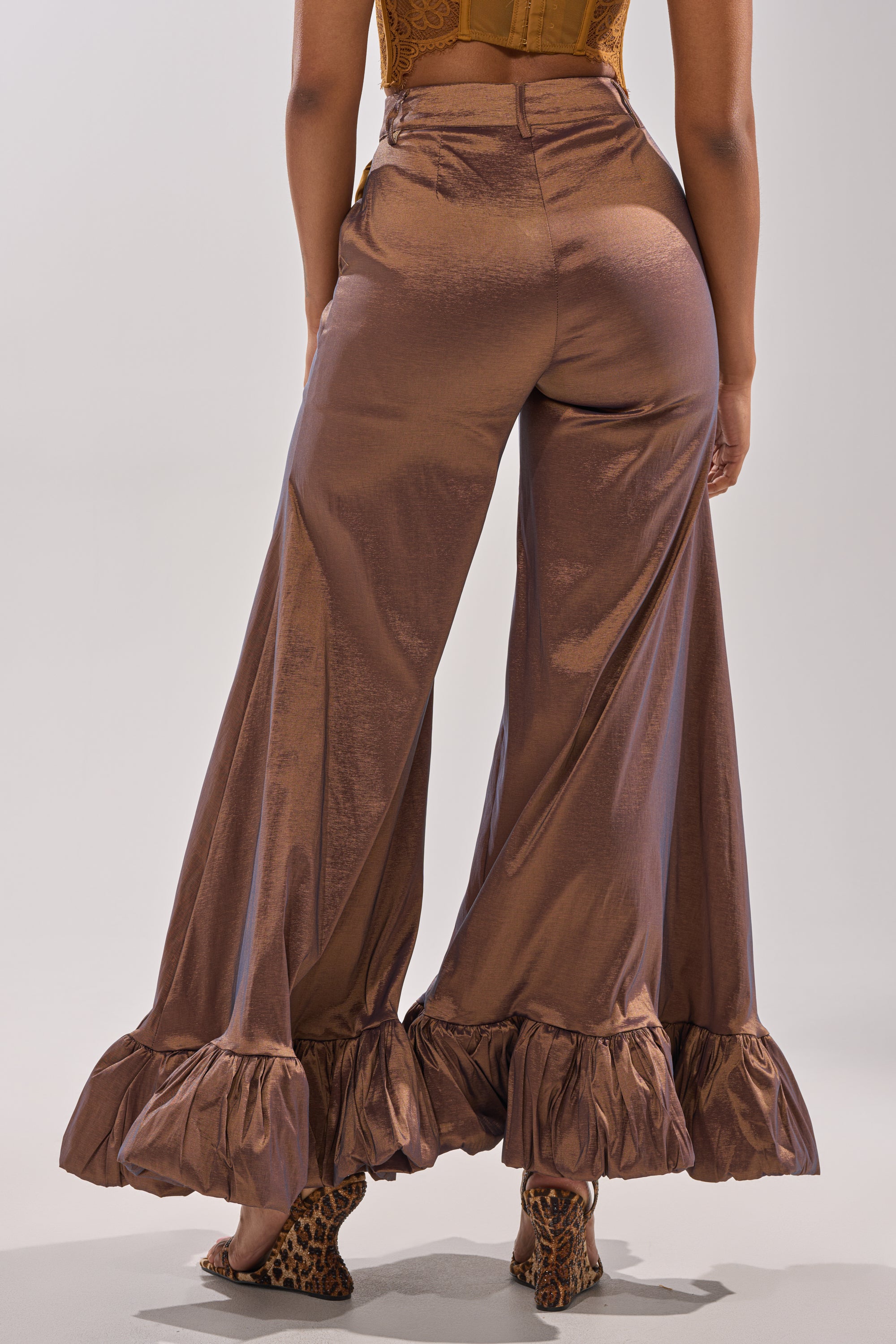 A person models the NOT TOO SIMPLE TROUSER—shiny brown metallic taffeta with wide legs and ruffled hems—paired with leopard-print heels, facing away from the camera against a plain background.