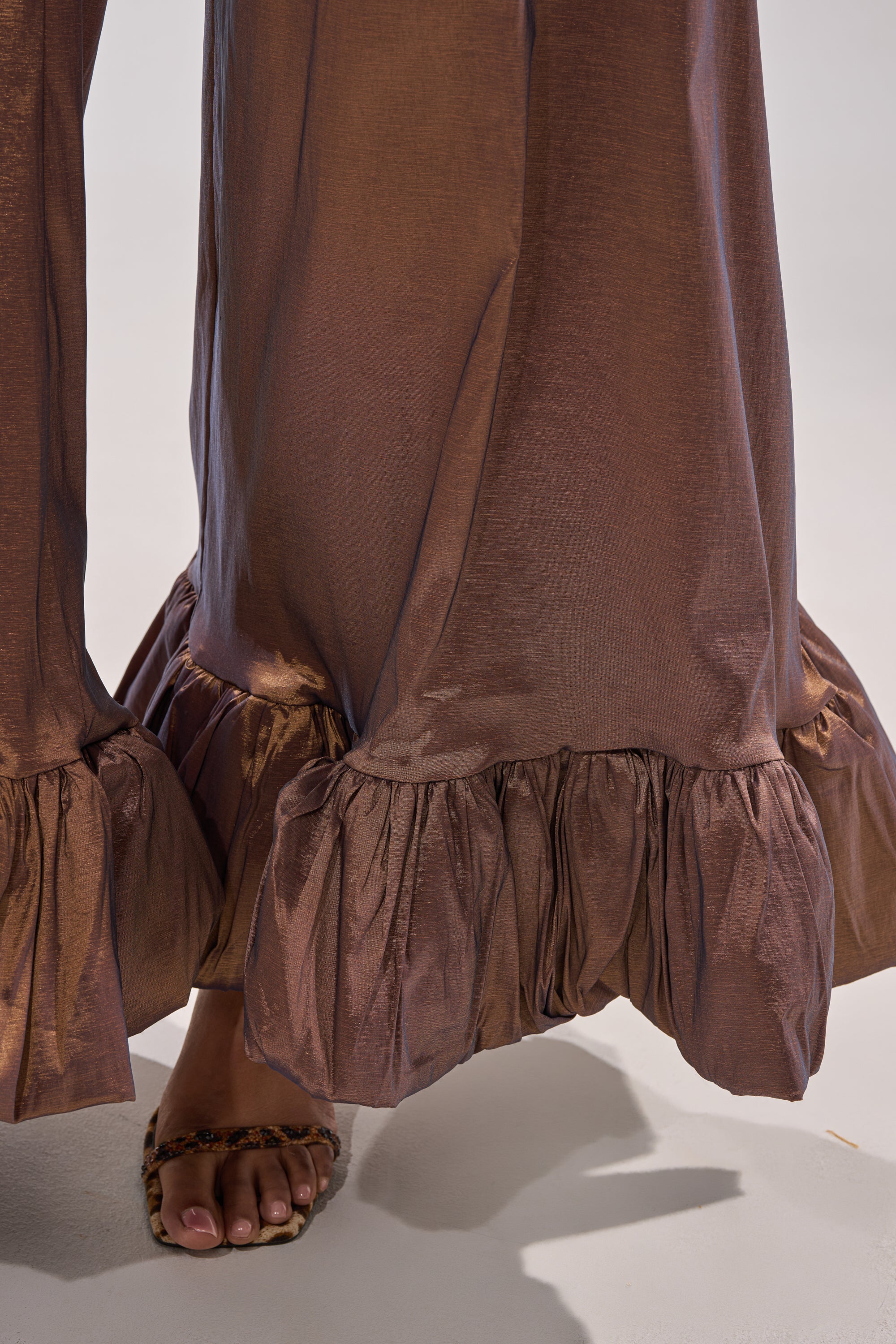 A close-up of a person wearing the NOT TOO SIMPLE TROUSER, metallic taffeta wide-leg pants with ruffled hems, paired with open-toe leopard print sandals on a light-colored floor.