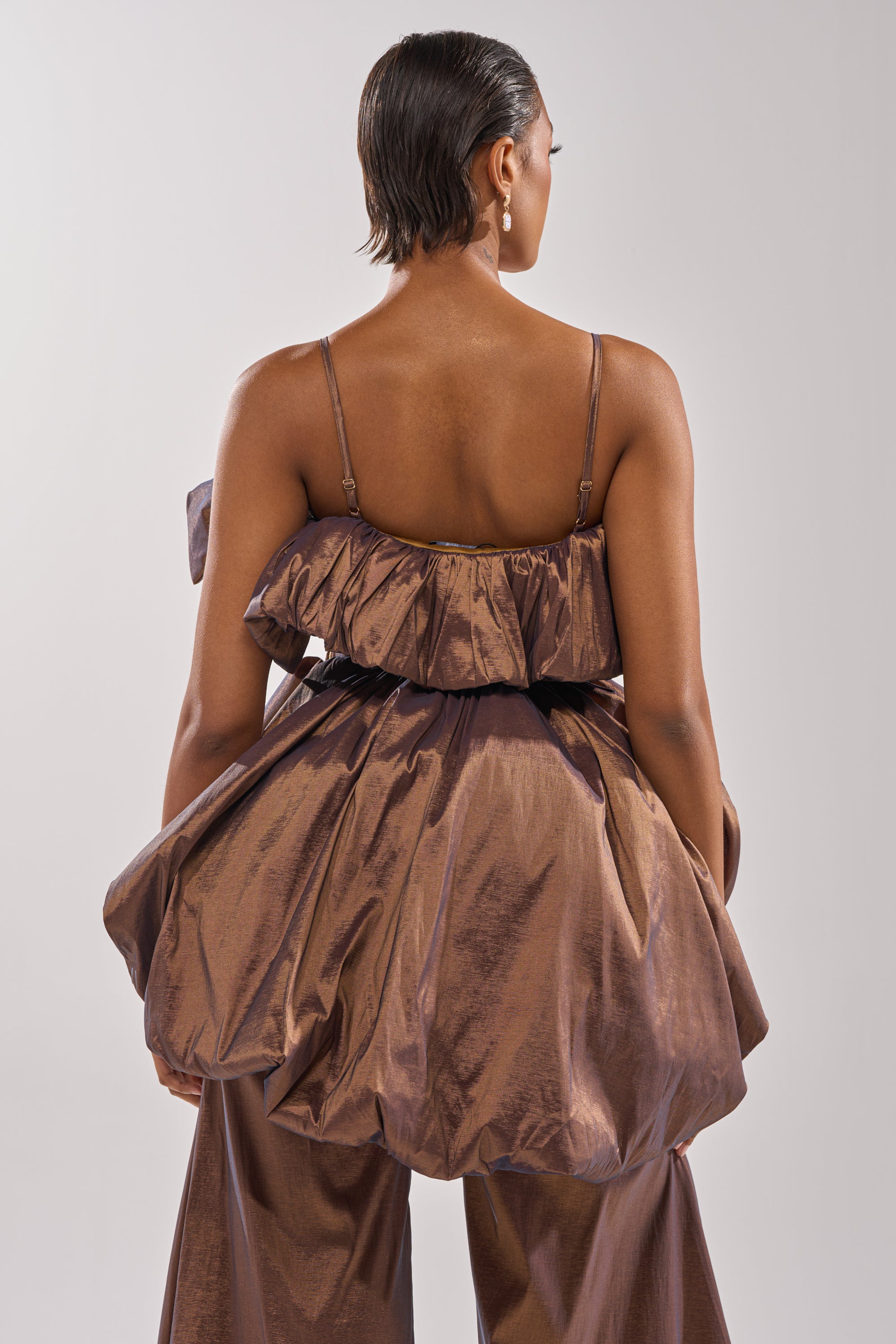 A woman with slicked-back short hair stands facing away, wearing the NOT TOO SIMPLE BLOUSE in shiny brown metallic taffeta with ruffles and thin straps, paired with wide matching pants against a plain light background.