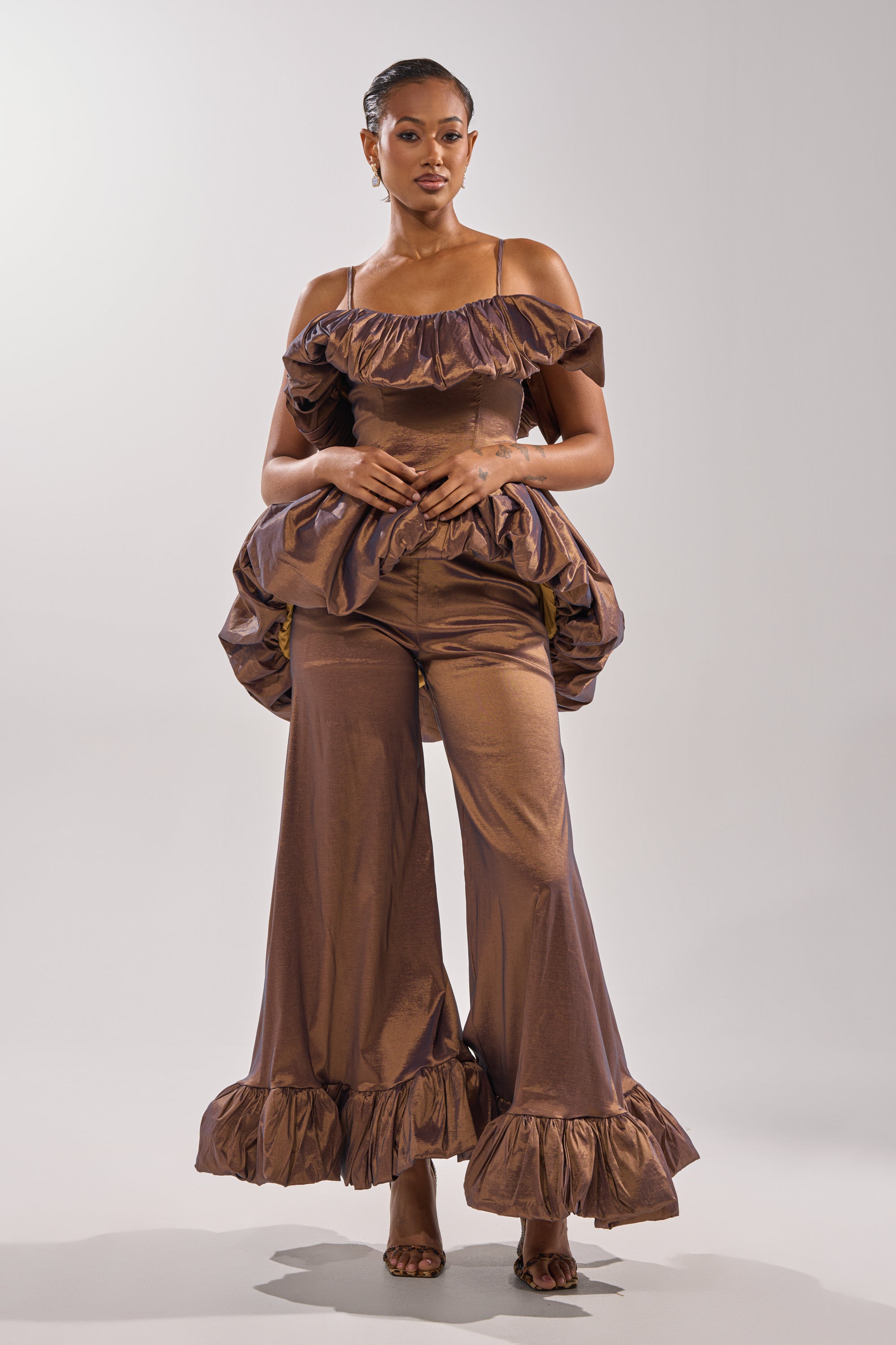 A woman poses confidently in the NOT TOO SIMPLE BLOUSE, a shiny bronze metallic taffeta top with ruffled off-the-shoulder sleeves and a peplum, styled with wide-leg pants featuring ruffled hems, hoop earrings, and open-toed heels.