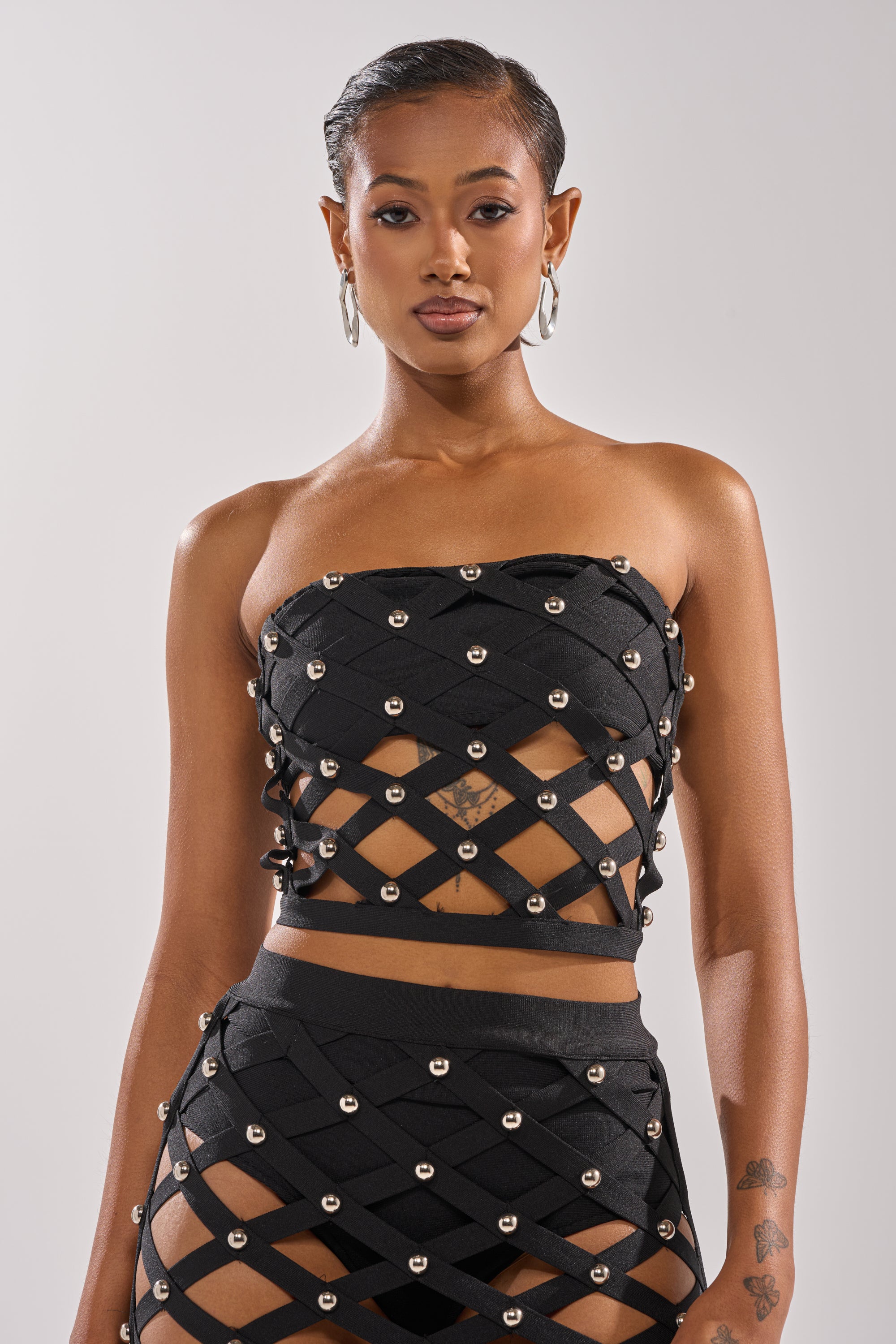 A woman with slicked-back hair models the CRAZY ENOUGH BANDEAU—a black bandeau top featuring a lattice of metal studs—paired with a matching skirt. She poses confidently against a simple, light background.