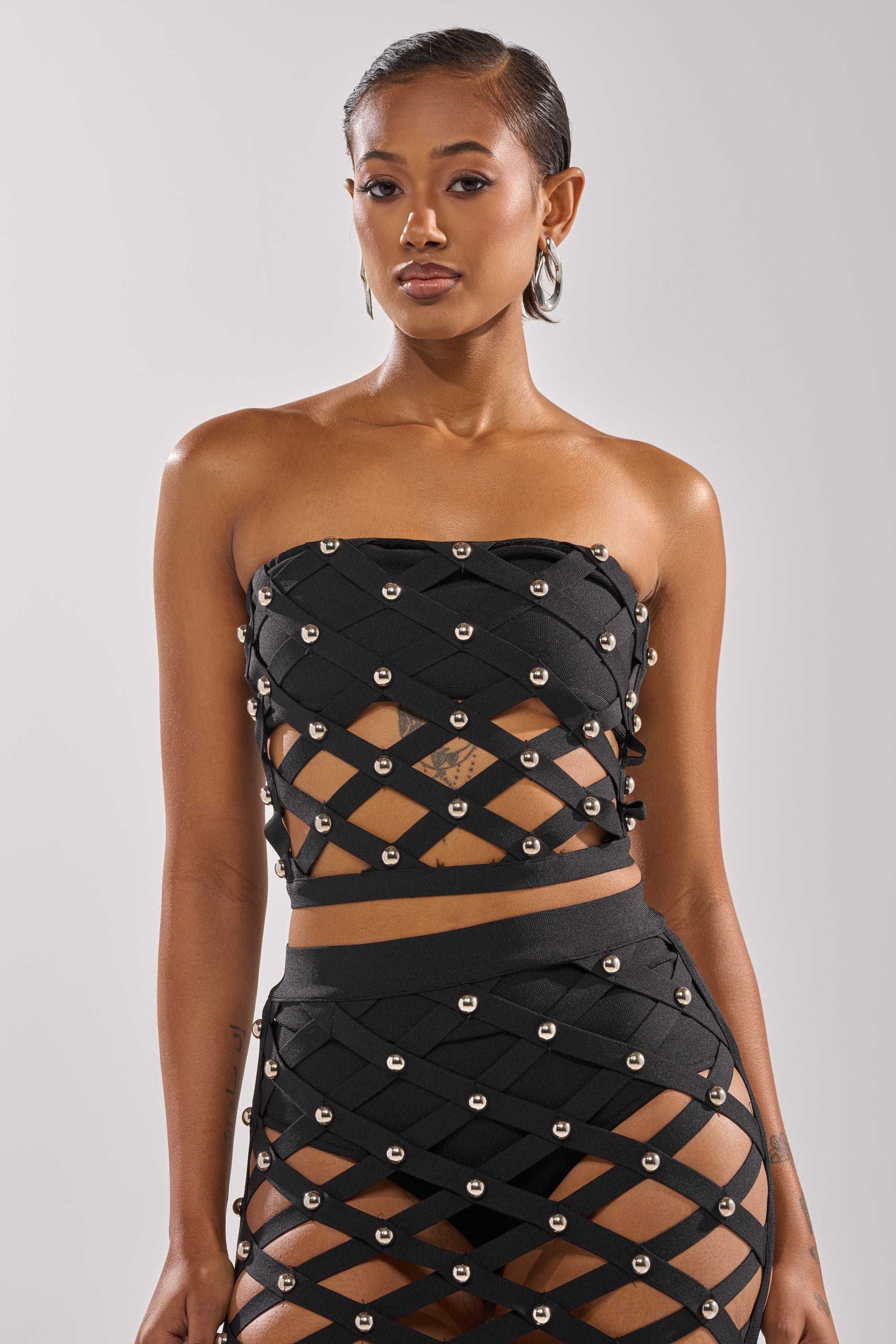 A woman wears the CRAZY ENOUGH BANDEAU—a black strapless bandeau top with a lattice design and metallic studs—paired with matching high-waisted bottoms, standing against a plain light background.