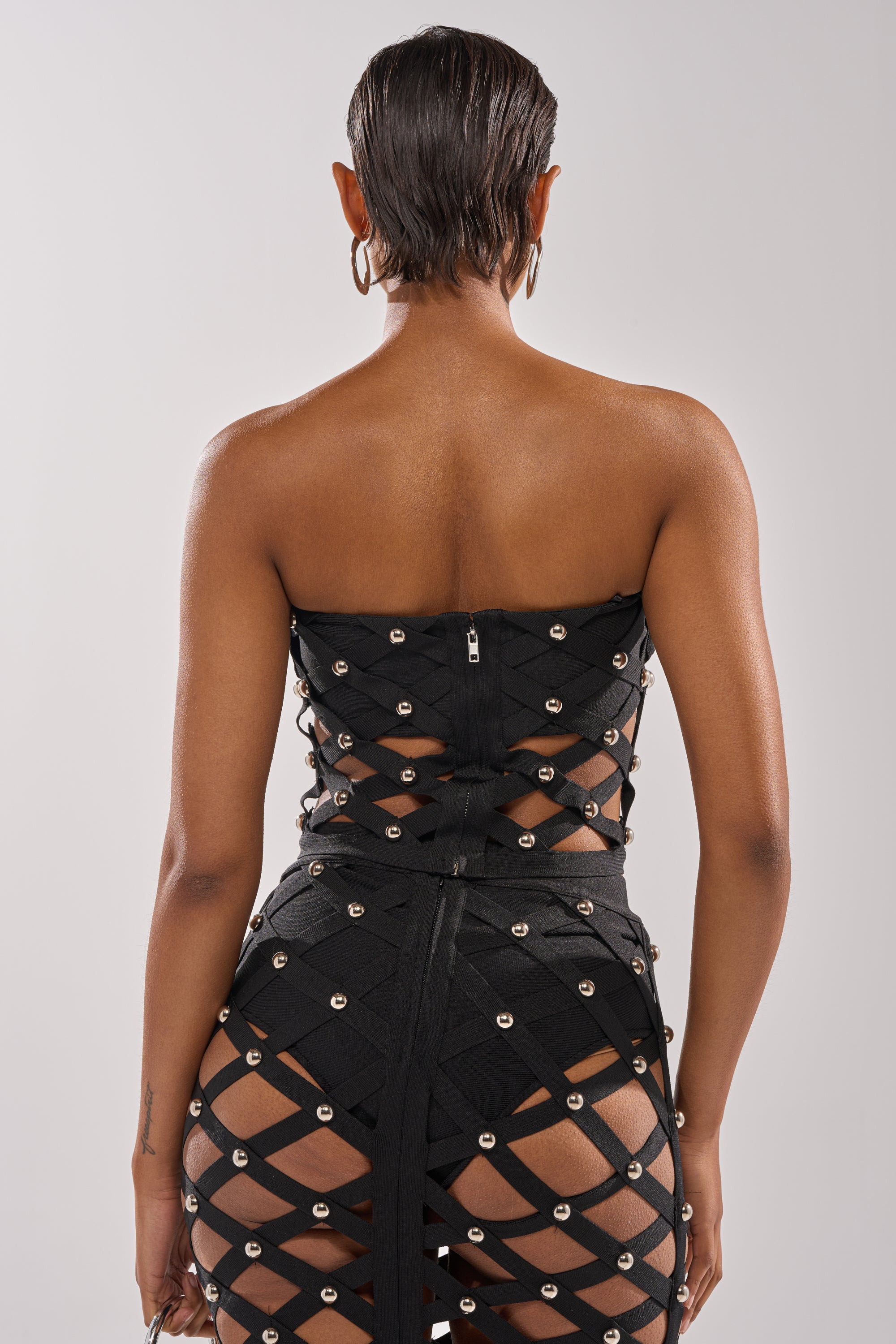 A person with short, slicked-back hair is seen from behind, wearing the CRAZY ENOUGH BANDEAU—a strapless black top featuring a lattice design with silver studs that reveals skin beneath its crisscrossed fabric.