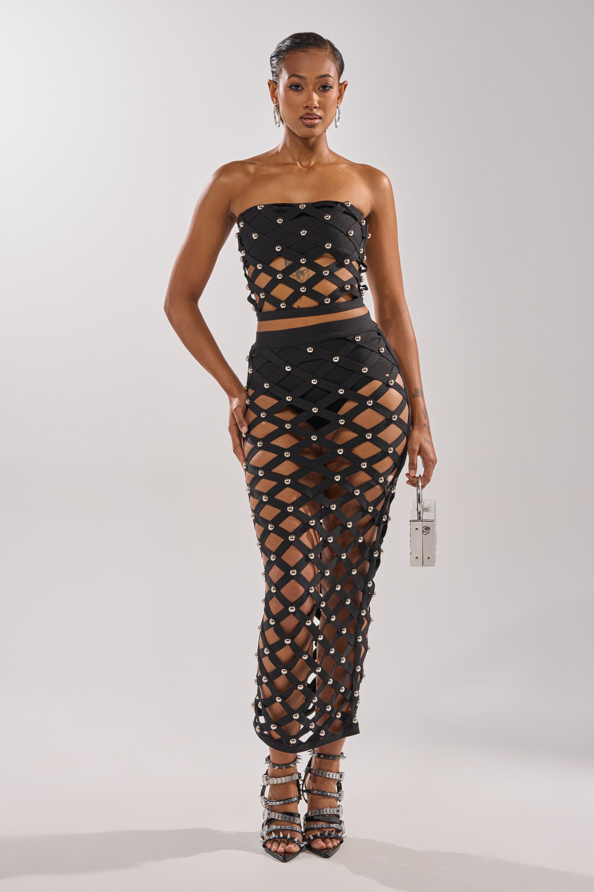 A woman poses against a simple backdrop in the black CRAZY ENOUGH MAXI SKIRT with crisscross cutouts and silver studs, paired with a matching strapless crop top. She accessorizes with a metallic purse and strappy high heels.