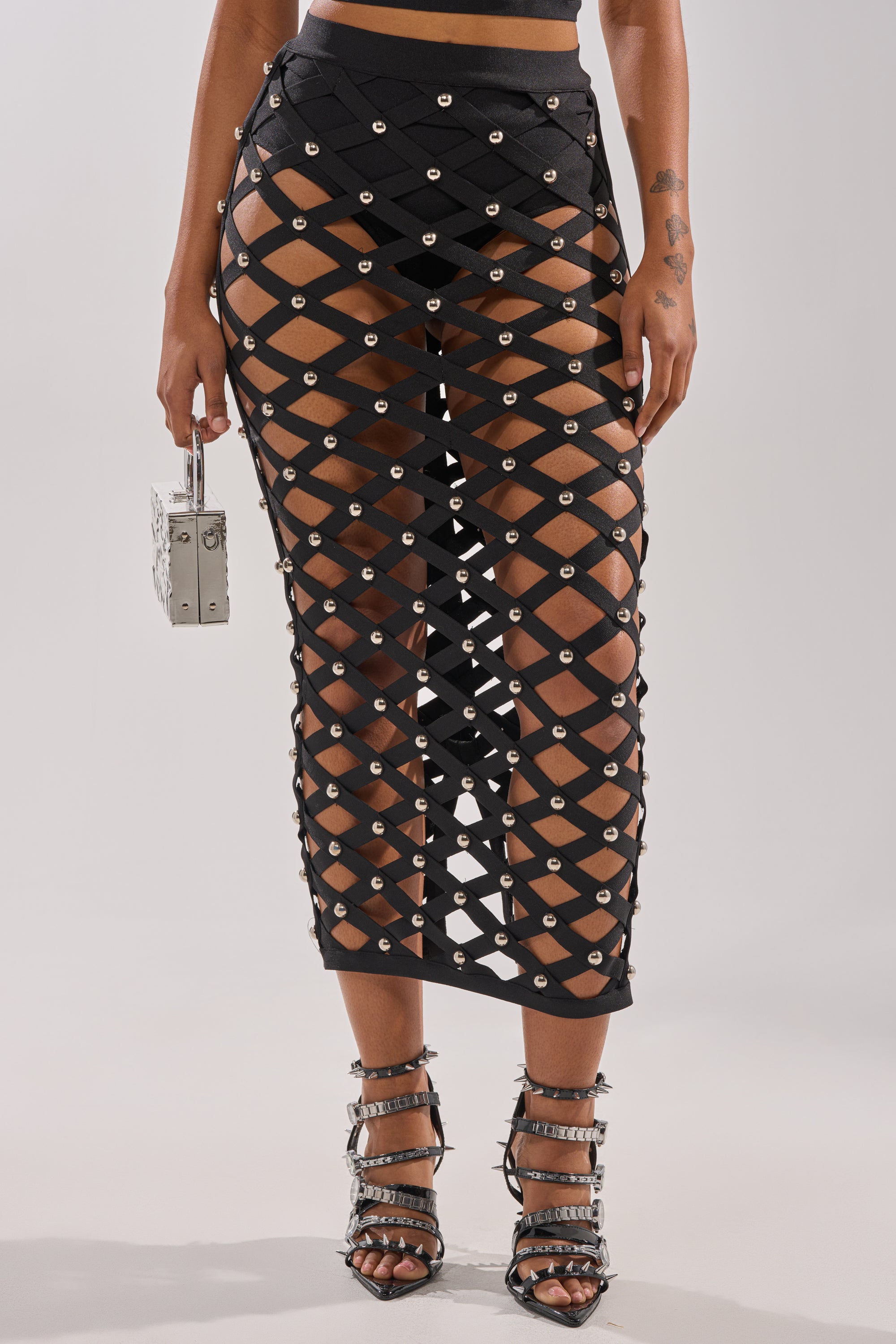 Wearing the CRAZY ENOUGH MAXI SKIRT—a black, cage-style silhouette with diamond cutouts and silver grommets—paired with strappy high heels and a metallic box purse.