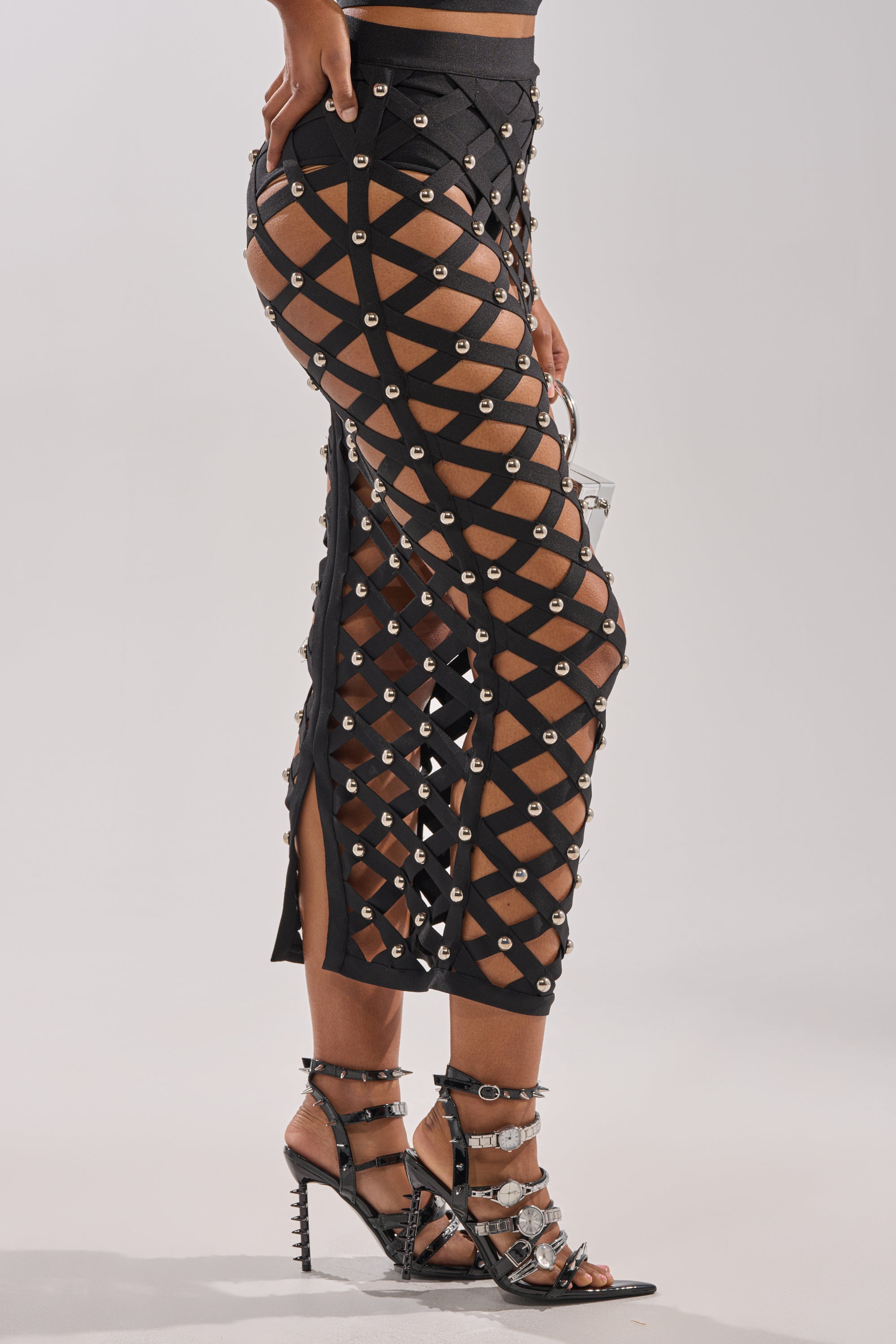 A person wears the CRAZY ENOUGH MAXI SKIRT, a black cage-style design with silver grommets that reveals skin beneath, styled with strappy black high-heeled sandals featuring silver details. The background is plain and light-colored.