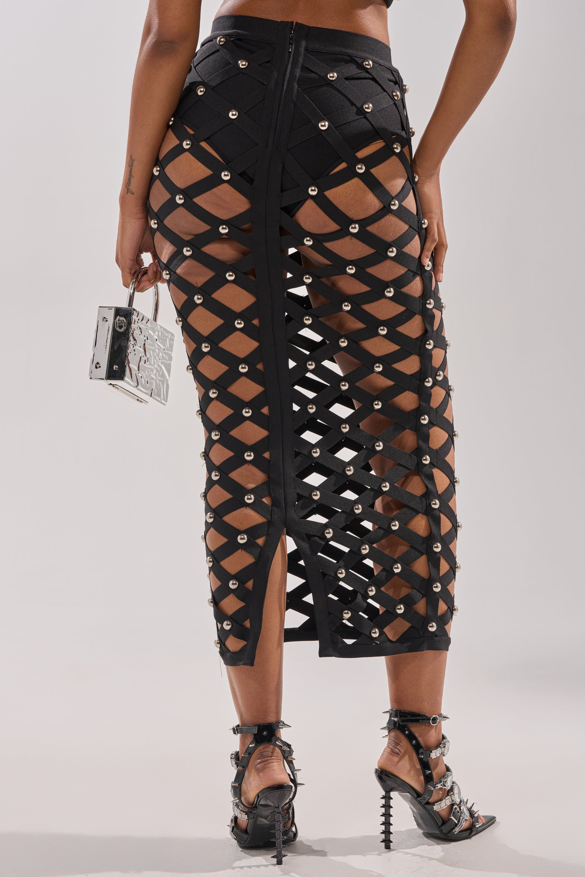 A person wearing the CRAZY ENOUGH MAXI SKIRT— a black cage-style midi with metal eyelets and a back slit— pairs it with black underwear, spiked high-heeled sandals, and holds a small metallic purse.