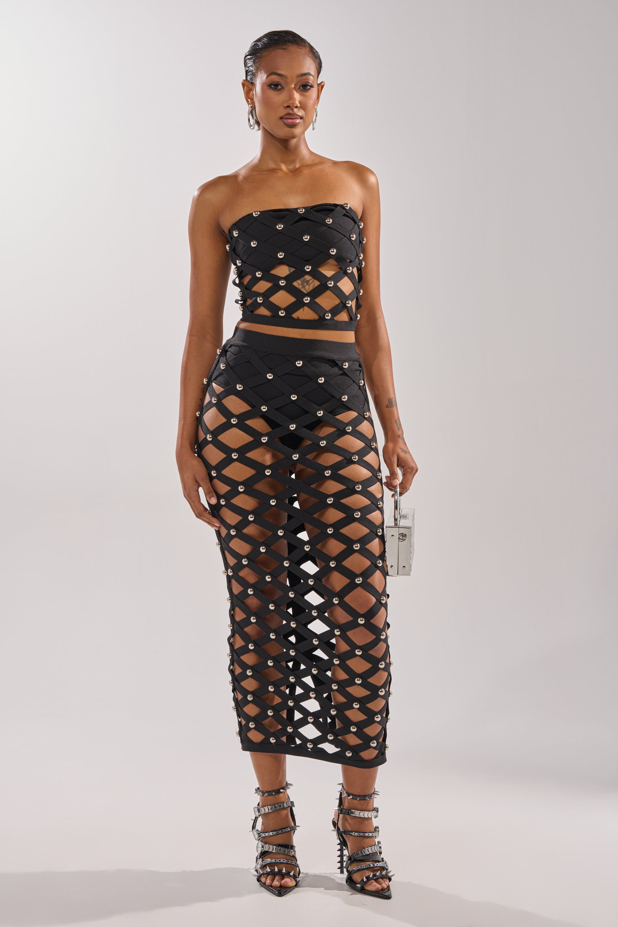 A woman poses in the CRAZY ENOUGH MAXI SKIRT, a high-waisted, cage-style black skirt with silver studs, paired with a strapless black crop top, strappy heels, and a metallic purse.