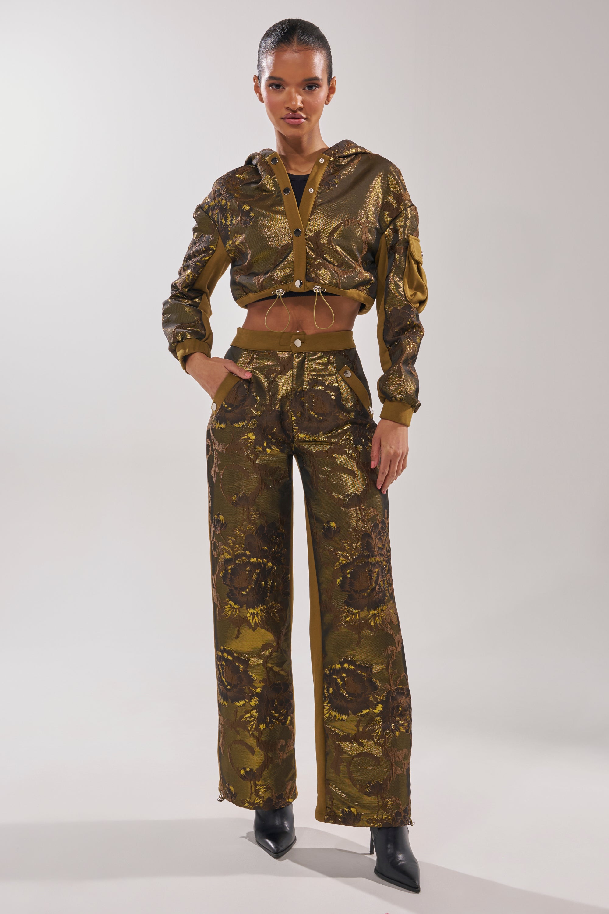 A woman stands confidently in a shiny olive green cropped jacket and the YOU KNOW IT BABY WIDE LEG JOGGER, which features a bold metallic floral pattern. She has her hands in her pockets and wears pointed black shoes. 0