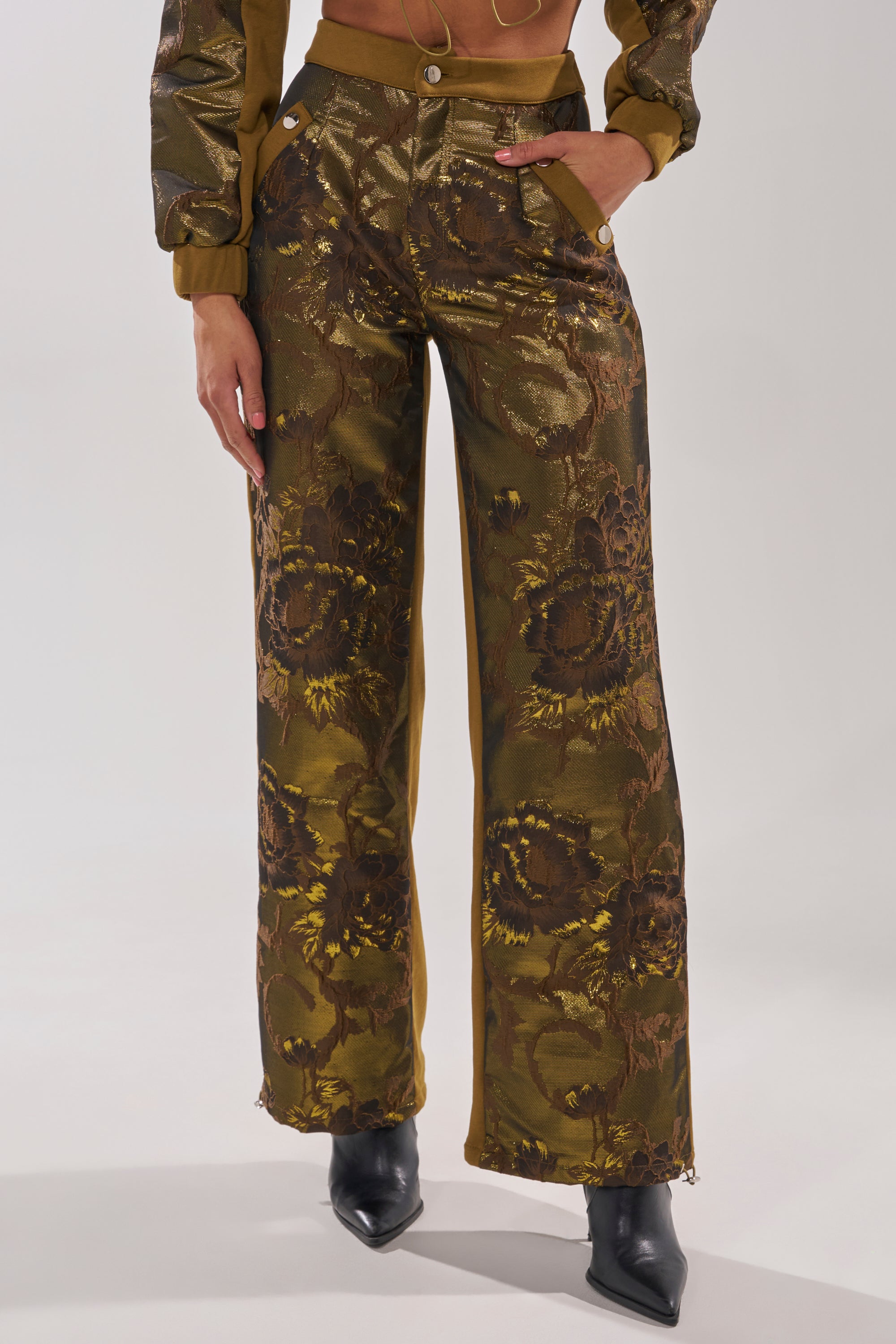 Wearing the YOU KNOW IT BABY WIDE LEG JOGGER—metallic brocade, high-waisted brown pants with a gold floral pattern—paired with a matching cropped jacket and pointed black boots. Only the lower body is visible against a plain background. 1