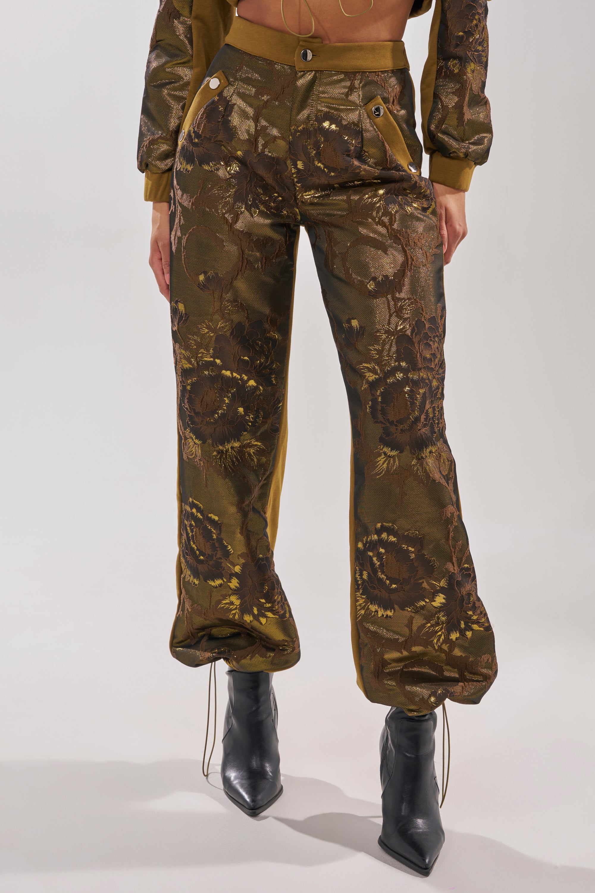 A person wearing the YOU KNOW IT BABY WIDE LEG JOGGER, shiny metallic brocade high-waisted pants with pleats, paired with black pointed-toe boots. The image is cropped from the chest down. 2