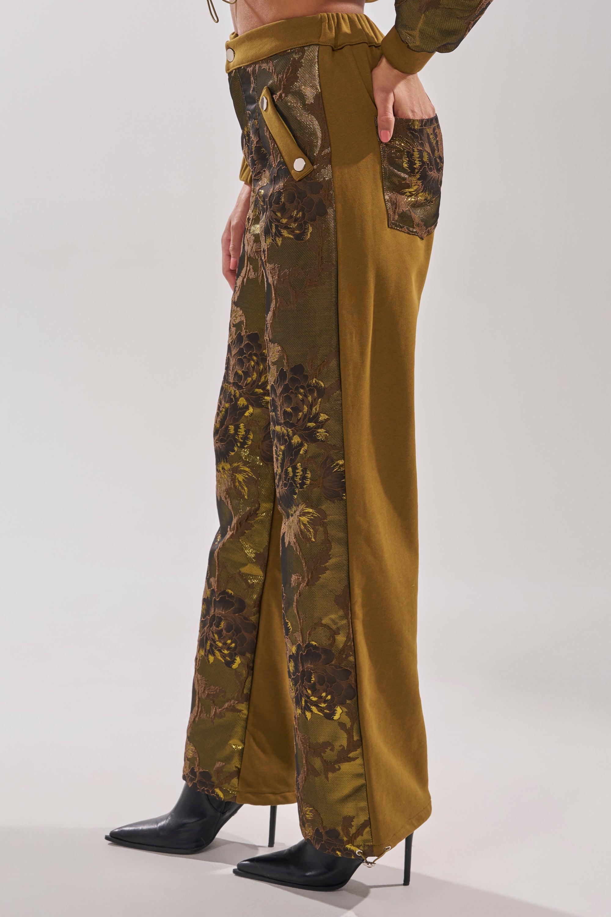 A person wearing the YOU KNOW IT BABY WIDE LEG JOGGER, featuring olive green and brown floral fabric panels with large decorative buttons, paired with black pointed-toe heeled boots. 4