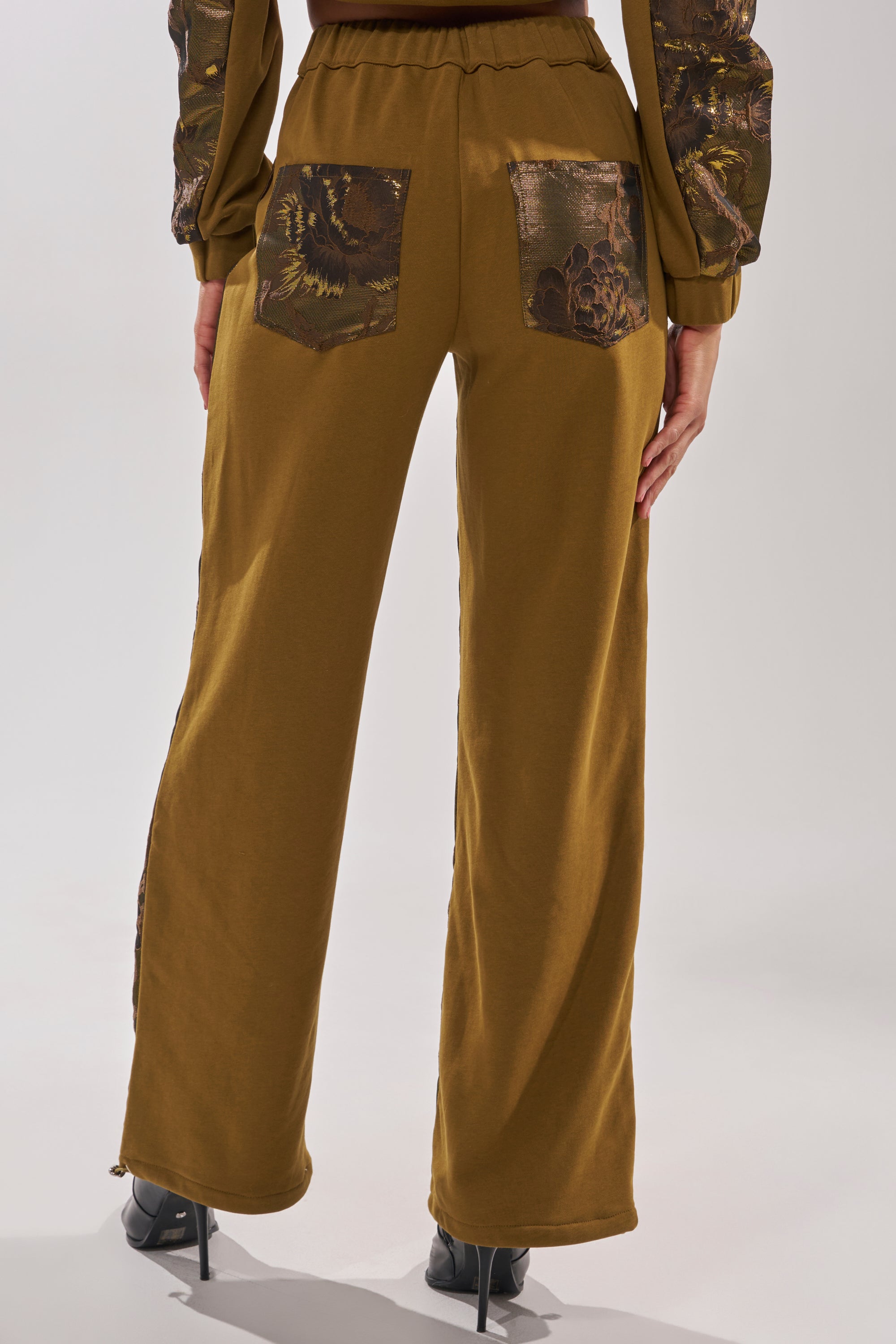 A person is shown from the back wearing the YOU KNOW IT BABY WIDE LEG JOGGER in olive green, featuring two patterned back pockets and a matching long-sleeve top, styled with black heeled shoes. 5