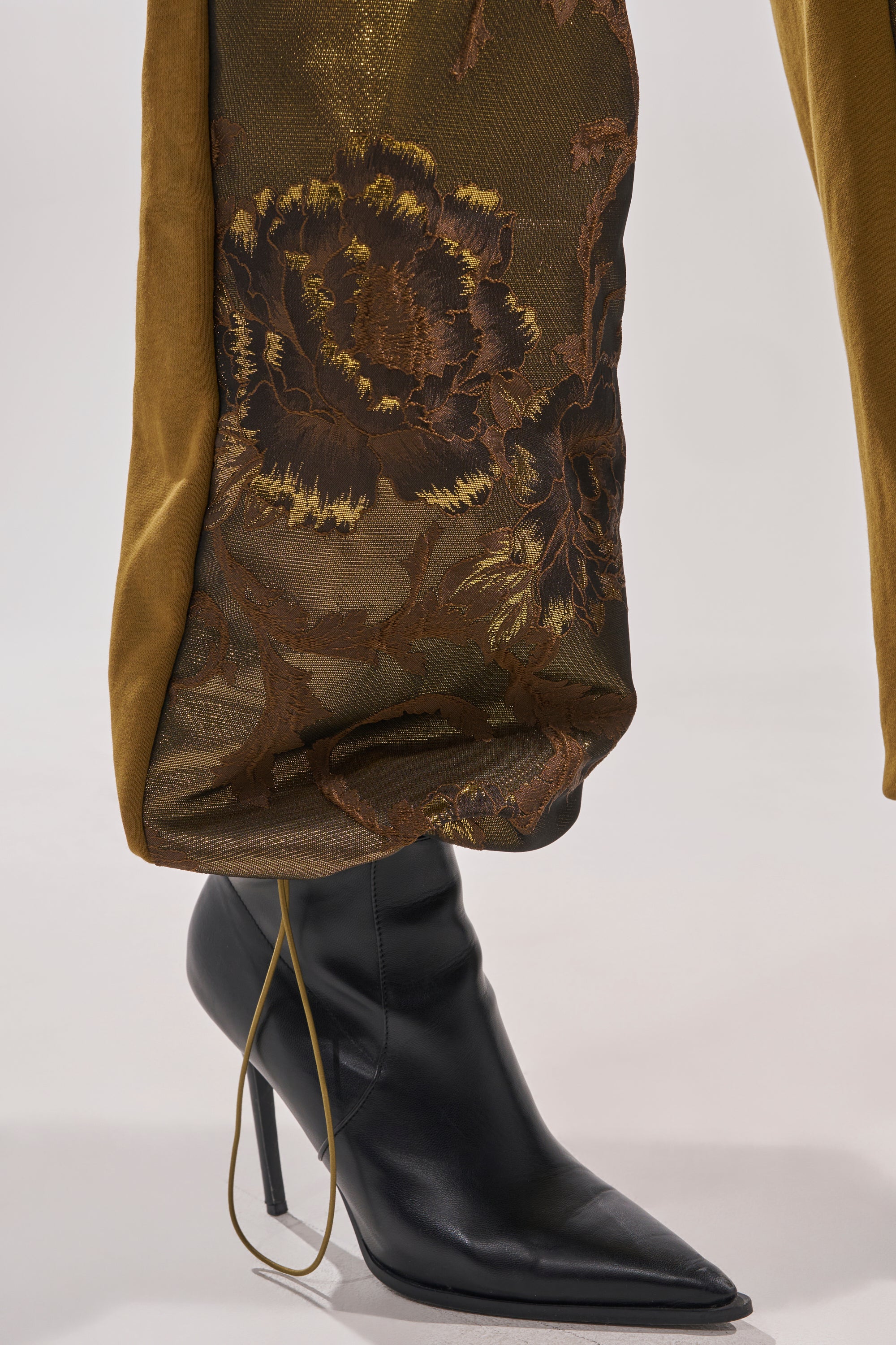 A close-up of a person wearing black pointed-toe heeled boots and YOU KNOW IT BABY WIDE LEG JOGGER pants in metallic brocade with a dark floral pattern, featuring long, high-rise legs slightly gathered at the ankle above the boots. 8