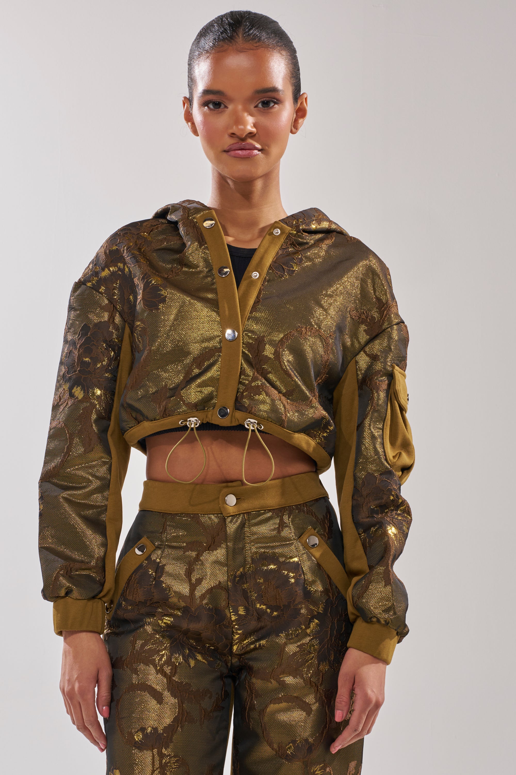 A woman models the YOU KNOW IT BABY CROP BUTTON DOWN, a shiny bronze and olive green patterned jacket with a hood and matching high-waisted pants, set against a plain light background. 1