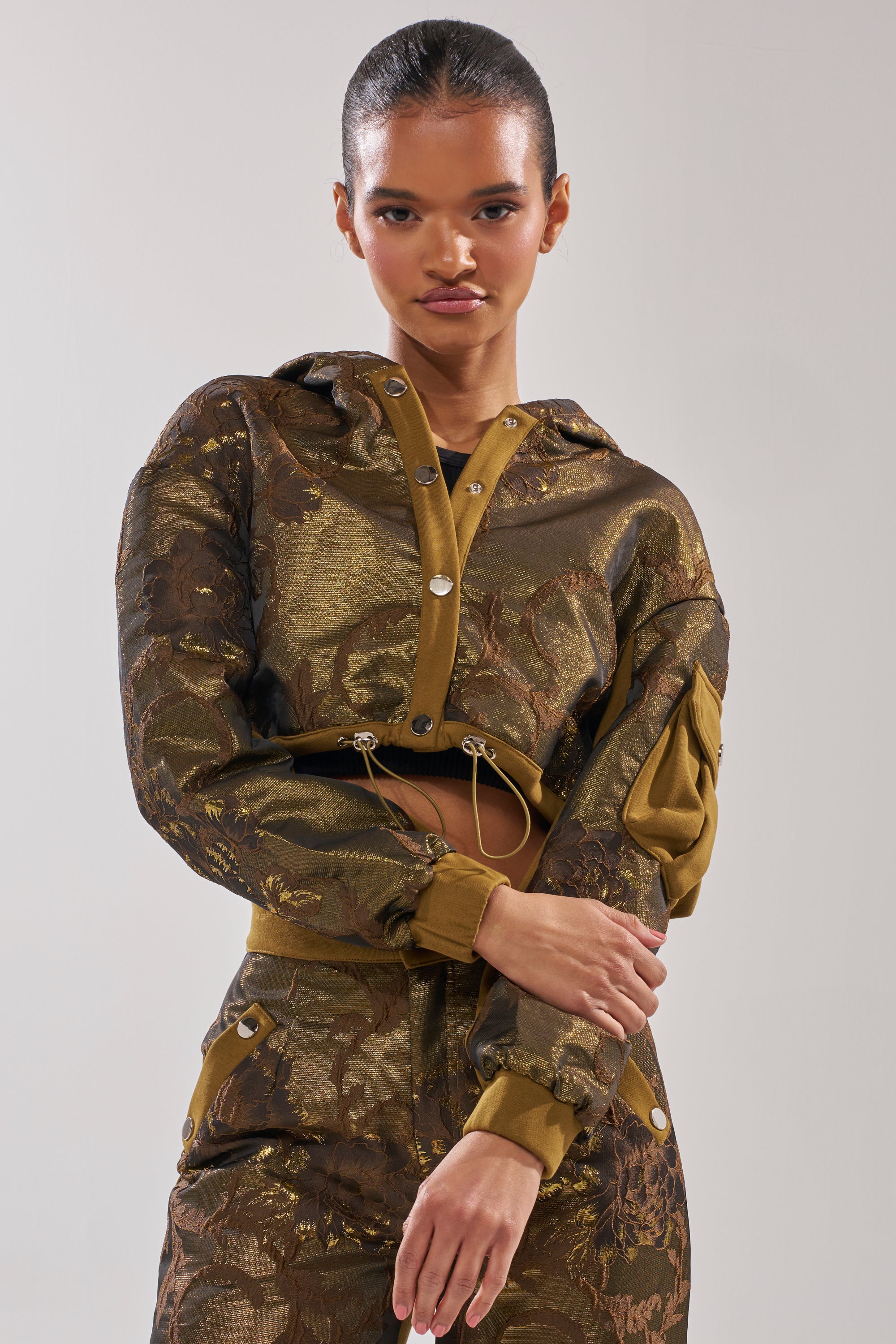 A woman models the YOU KNOW IT BABY CROP BUTTON DOWN, a metallic gold and brown patterned hooded jacket with matching pants and a floral brocade panel, posing with her arms crossed against a plain light background. 2