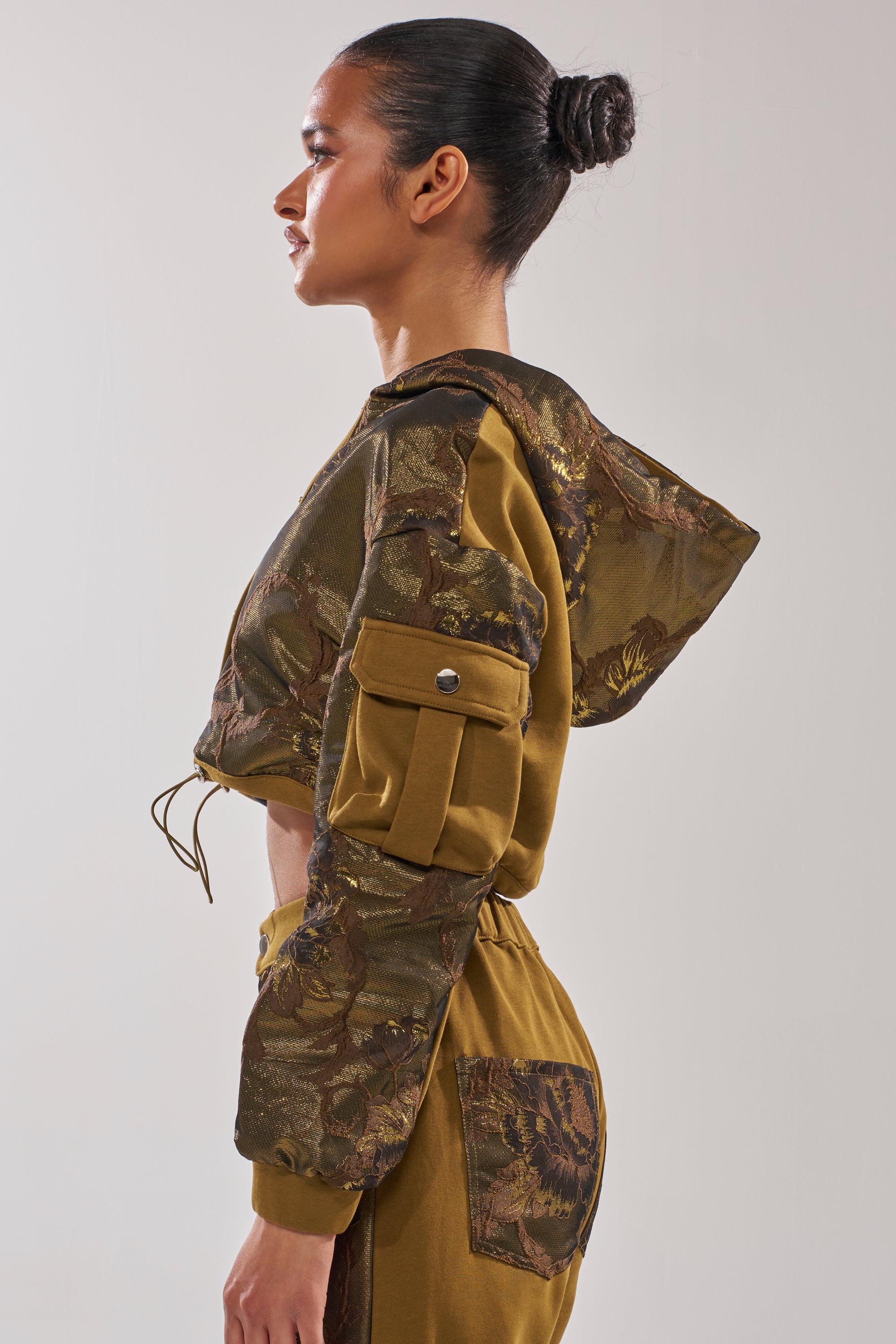 A woman stands in profile wearing the YOU KNOW IT BABY CROP BUTTON DOWN, a streetwear-inspired cropped hooded jacket with a brown and gold metallic leaf pattern, cargo sleeve pocket, and paired with matching high-waisted pants. Her hair is styled in a neat bun. 3