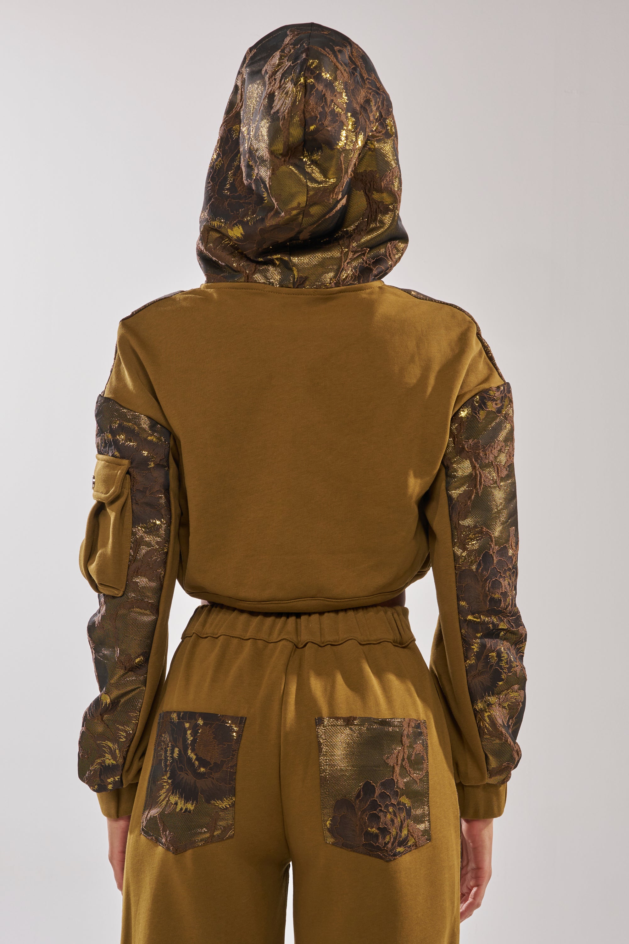 A person faces away, wearing the YOU KNOW IT BABY CROP BUTTON DOWN in olive green brocade with a shiny floral-patterned hood, sleeves, and pockets. The matching pants have the same bold floral design on the back pockets. 4