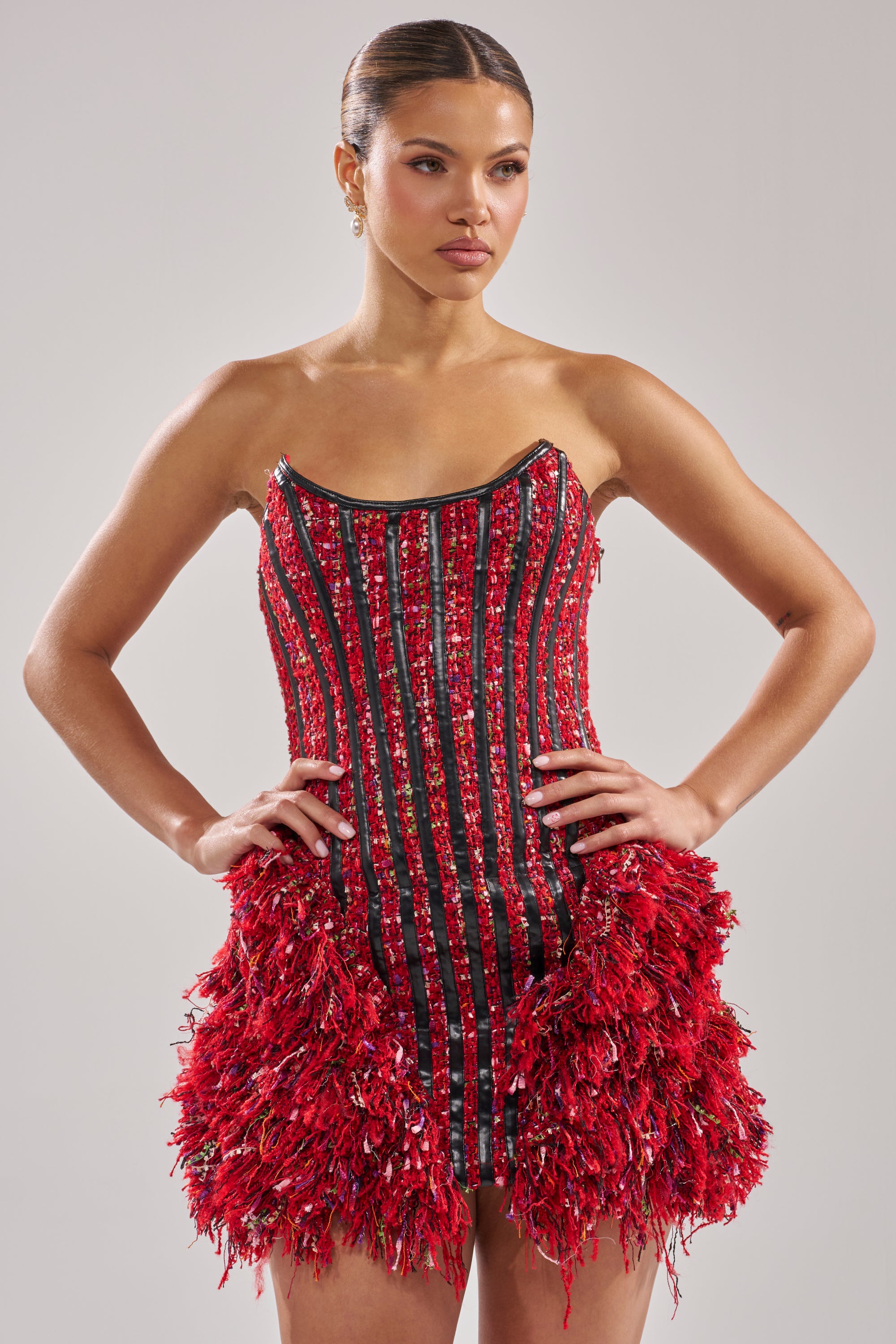 A woman stands with her hands on her hips, wearing the LEAH TWEED MINI DRESS—a striking mini with vertical stripes, a corset bodice, and a textured fringed skirt. Her sleek bun and neutral expression complete this perfect holiday dress look. 0