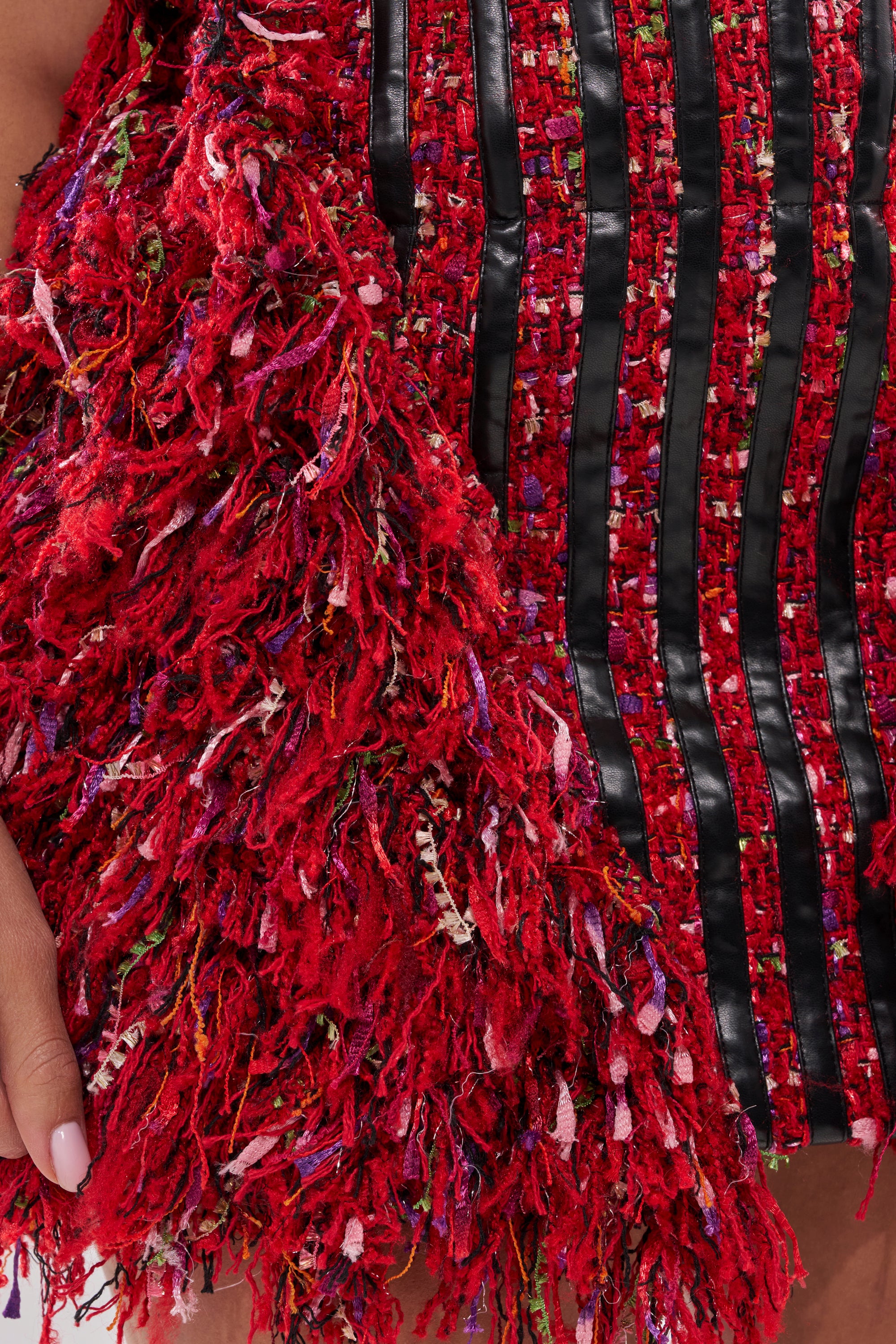 Close-up of the LEAH TWEED MINI DRESS: red holiday style with shaggy, fringed fabric on one side and vertical black stripes with a woven red pattern on the other. A hand with light pink nail polish appears on the left. 5