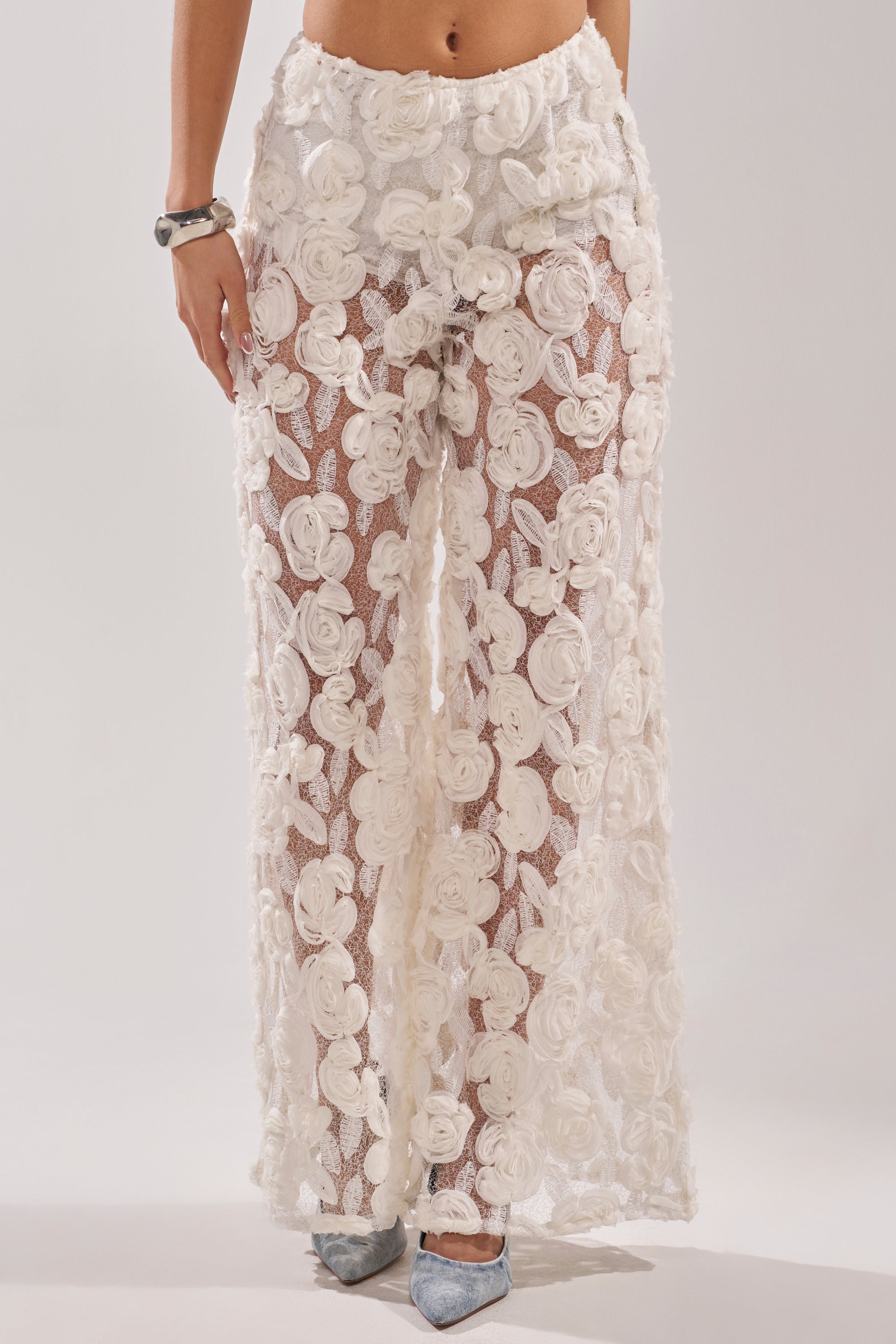 A person wears the CRYSTAL PALAZZO PANT—sheer white pants with large 3D floral appliqués—paired with light blue pointed-toe shoes and a silver bracelet, set against a plain, neutral background.