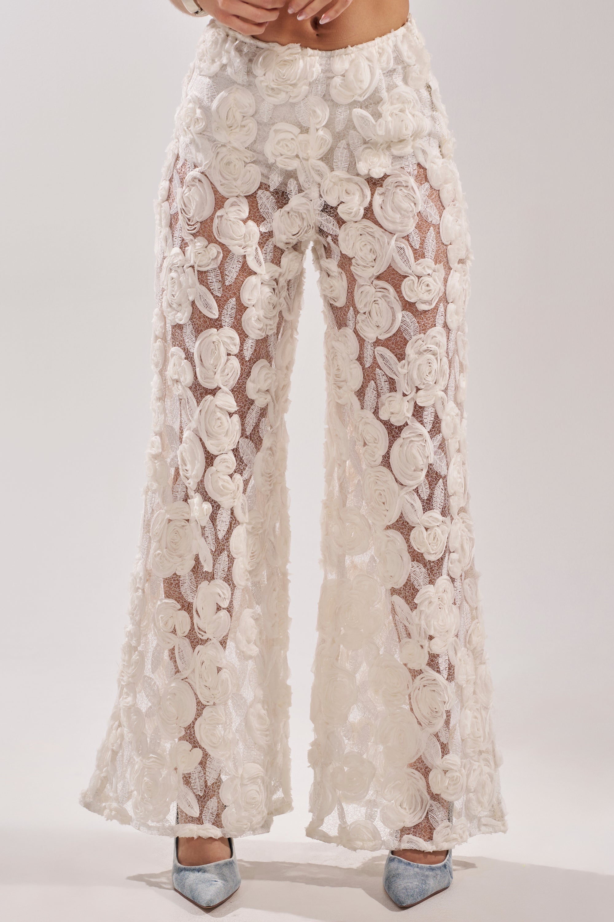 A person wears the CRYSTAL PALAZZO PANT—high-waisted, wide-leg, 3D floral sheer pants with delicate rosette appliqué—paired with light blue pointed-toe heels against a plain, light-colored background.