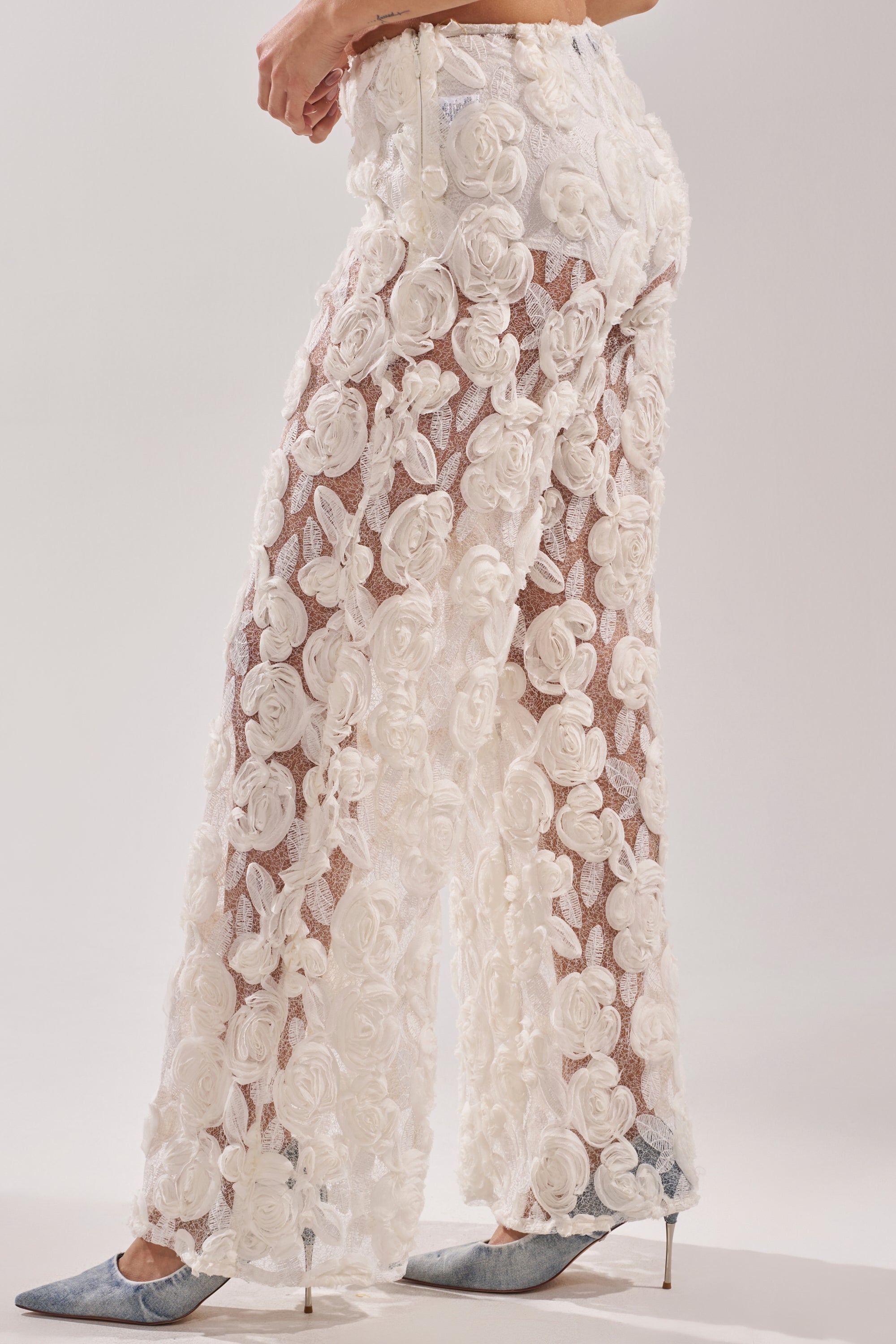 A person wears CRYSTAL PALAZZO PANT sheer wide-leg pants with large 3D floral appliqués and blue pointed-toe heels, shown waist down against a plain background.