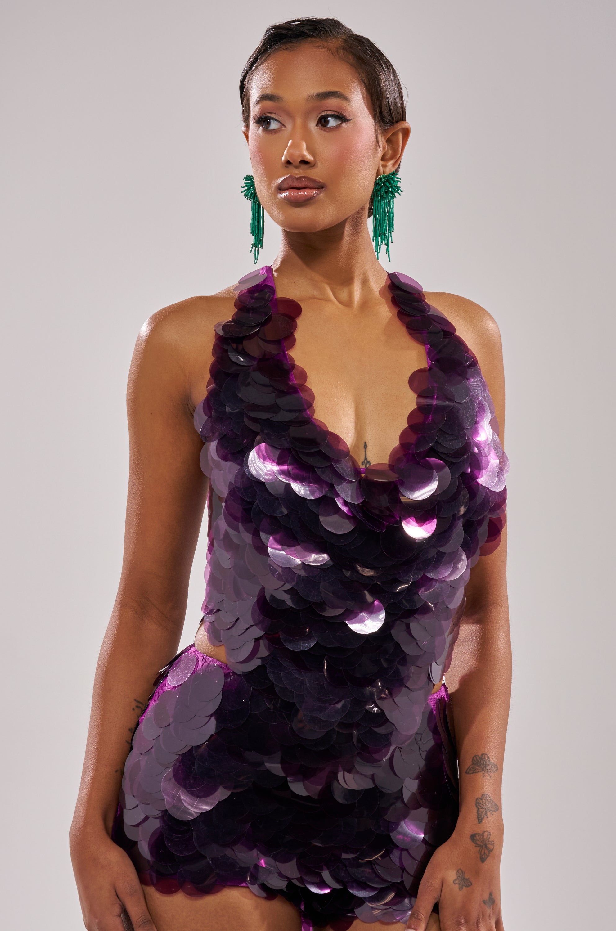 A woman wearing the LOVE BOMB SEQUIN BLOUSE, a sleeveless purple halter-style top, stands confidently against a plain background. She has short slicked-back hair, green statement earrings, and tattoos on her right forearm.