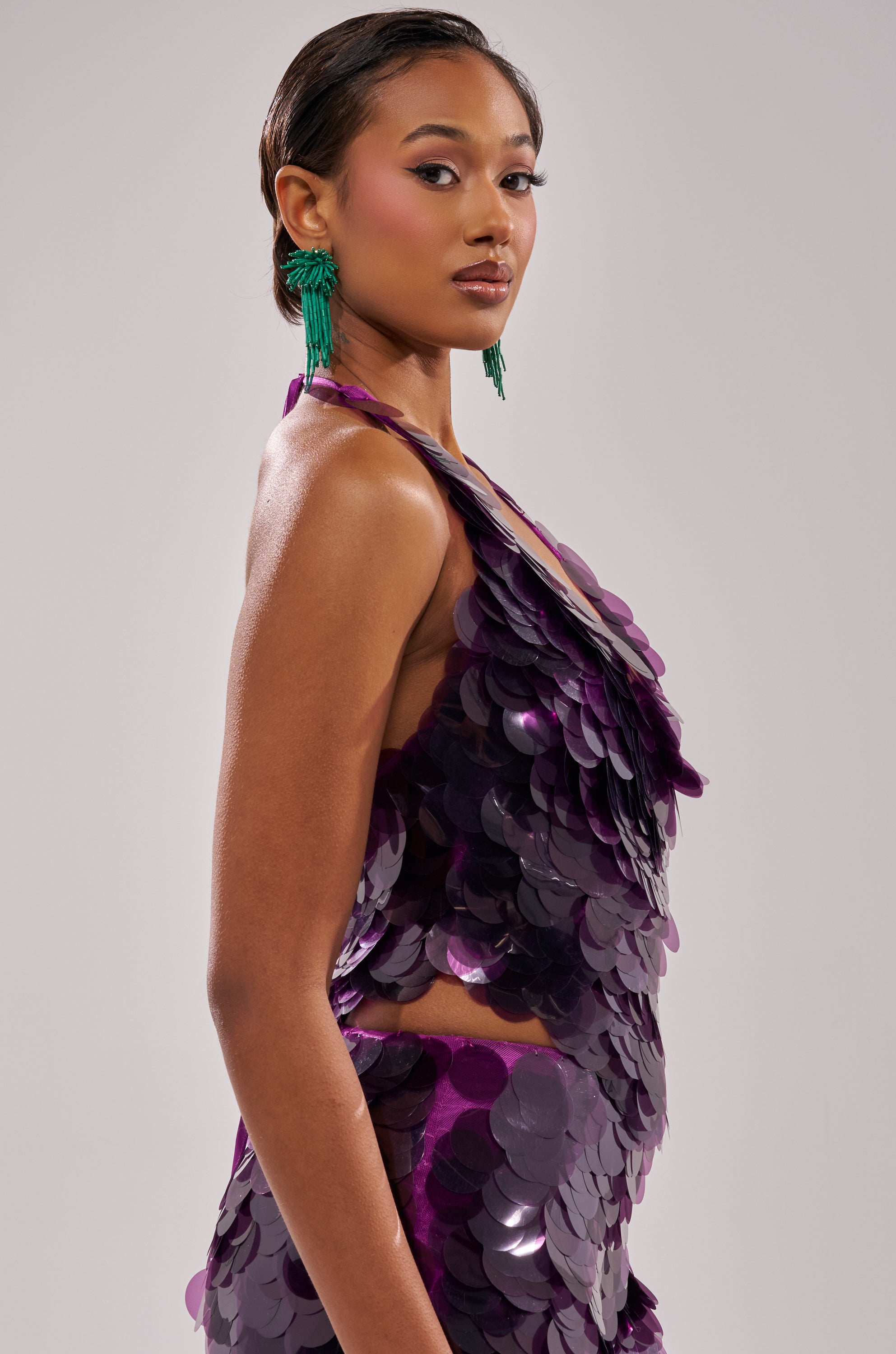 A woman models the LOVE BOMB SEQUIN BLOUSE, featuring halter styling and large, shiny purple sequins. She wears slicked-back hair, green statement earrings, and bold makeup against a plain light background.